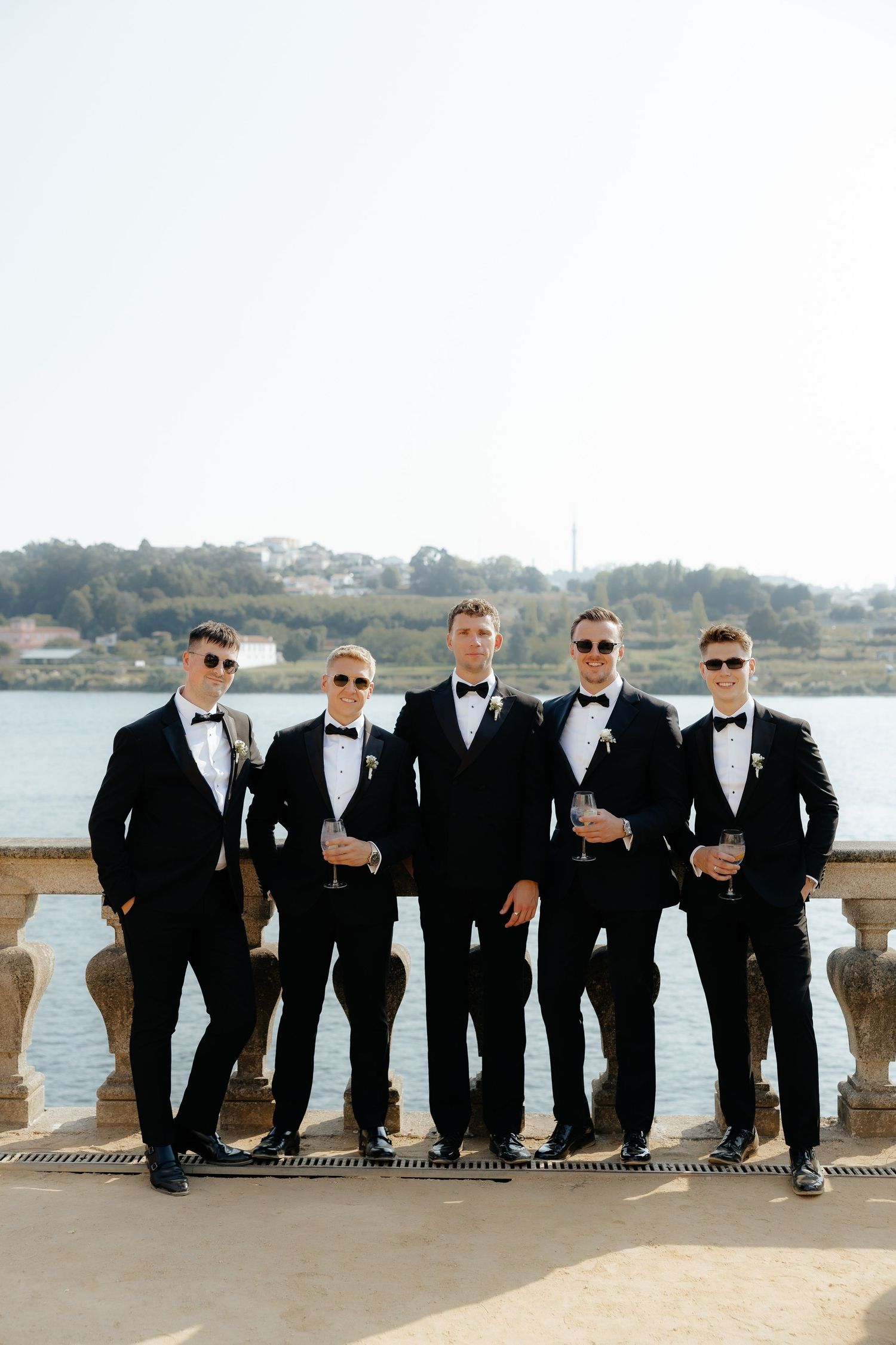 Groomsmen in black tuxedos lean casually against a stone balustrade overlooking a waterfront view.