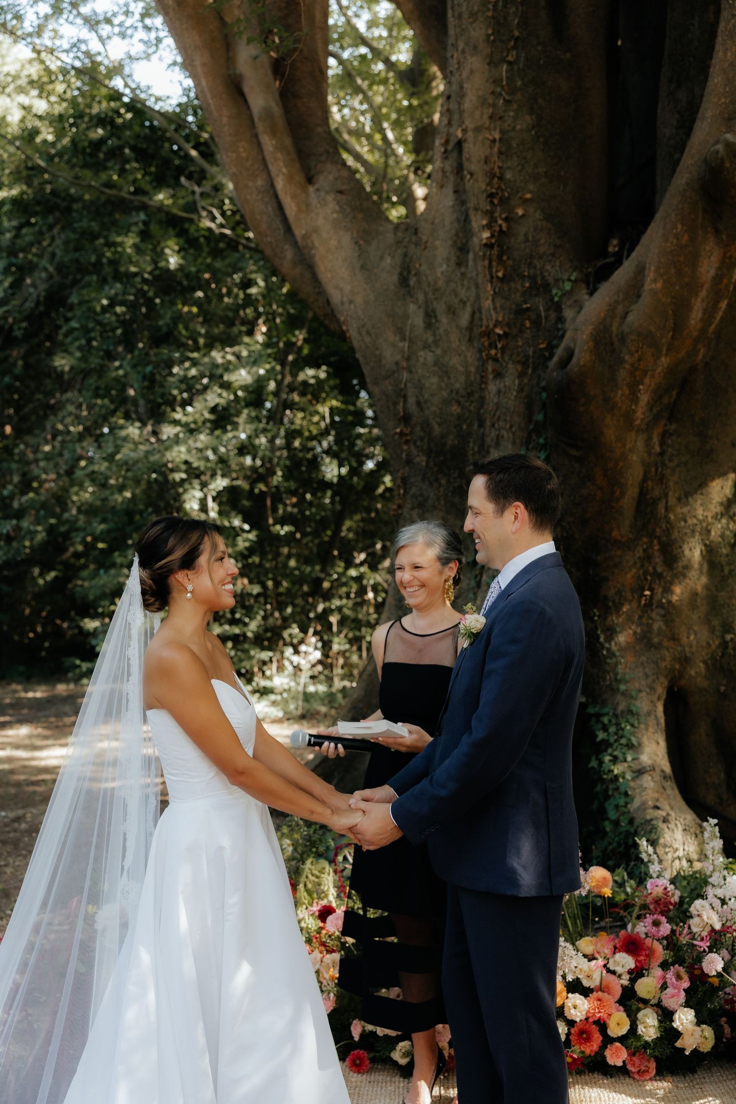 A wedding ceremony takes place under large trees with pink and orange floral arrangements.