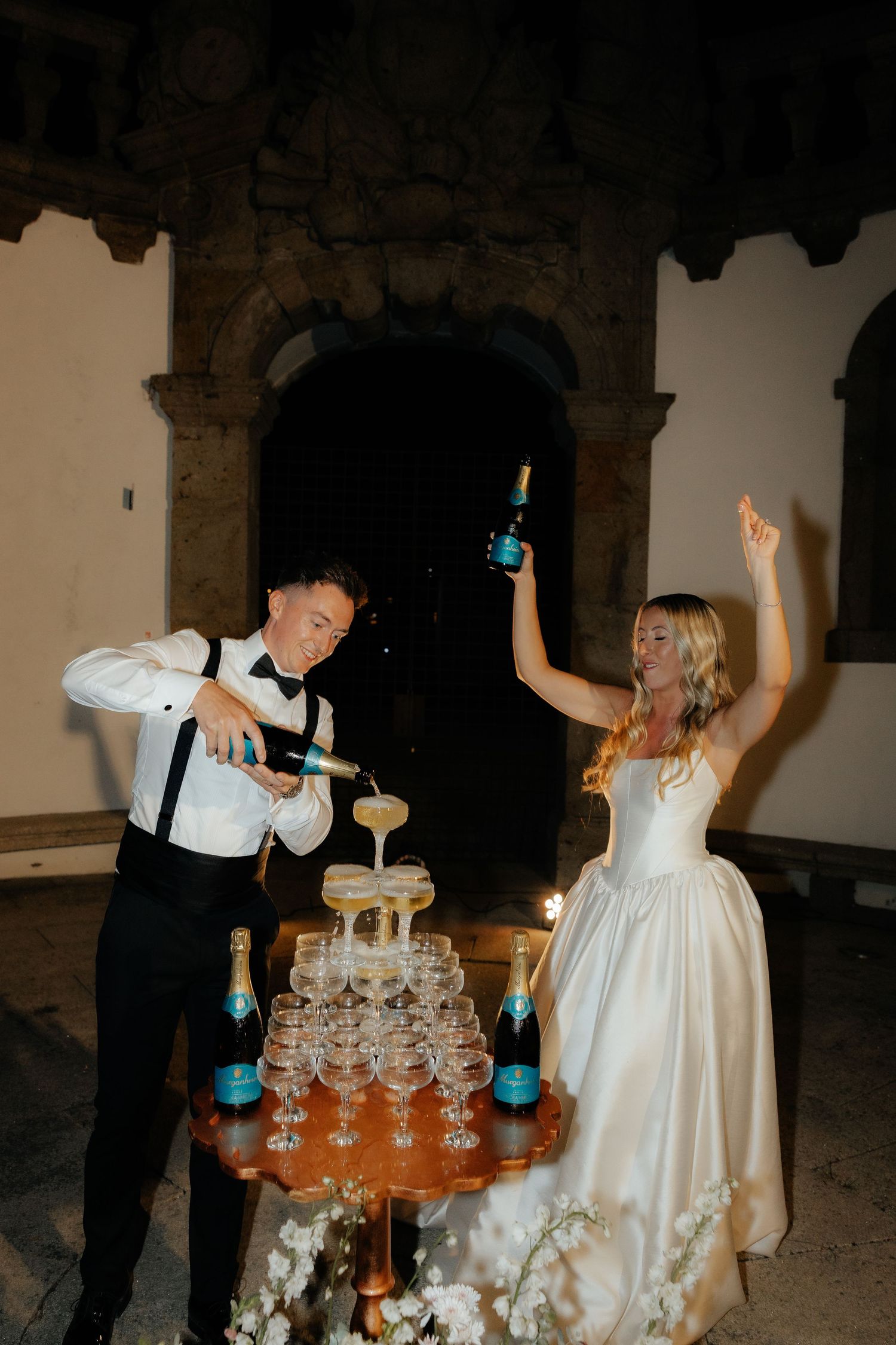 Newlyweds laughing while pouring drinks into a champagne tower at their reception.