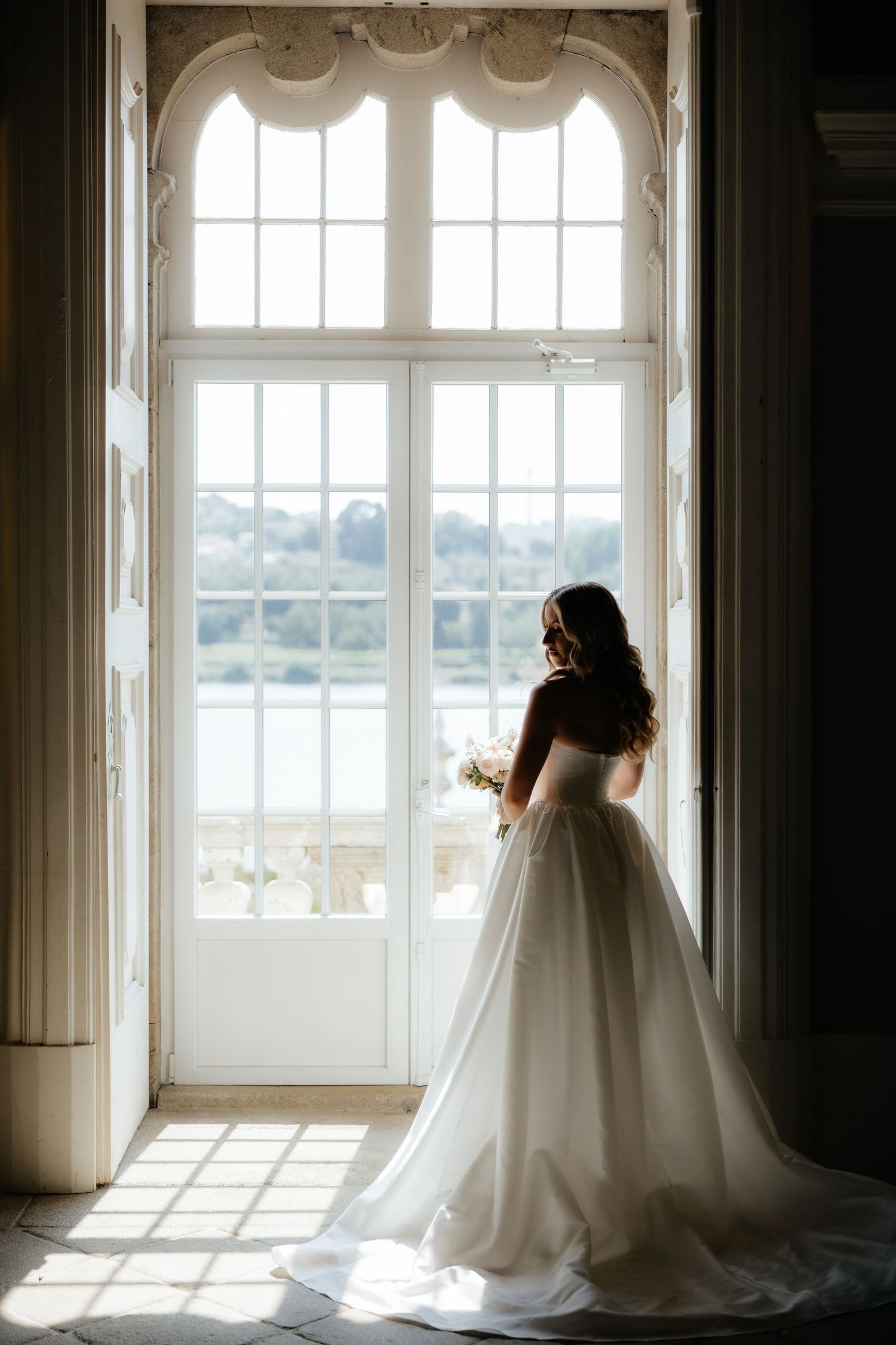 A bride in a flowing white wedding gown stands silhouetted against bright sunlit windows in an elegant room.