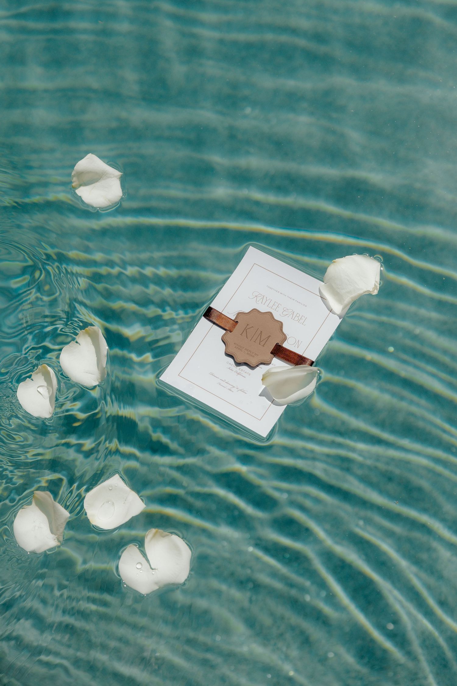 Wedding invitation with leather monogram tag floating in turquoise pool water with white rose petals.