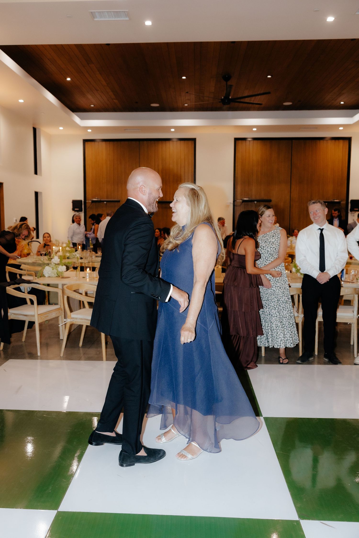 Groom dances with a woman in a navy blue gown on a white checkered dance floor during wedding reception.
