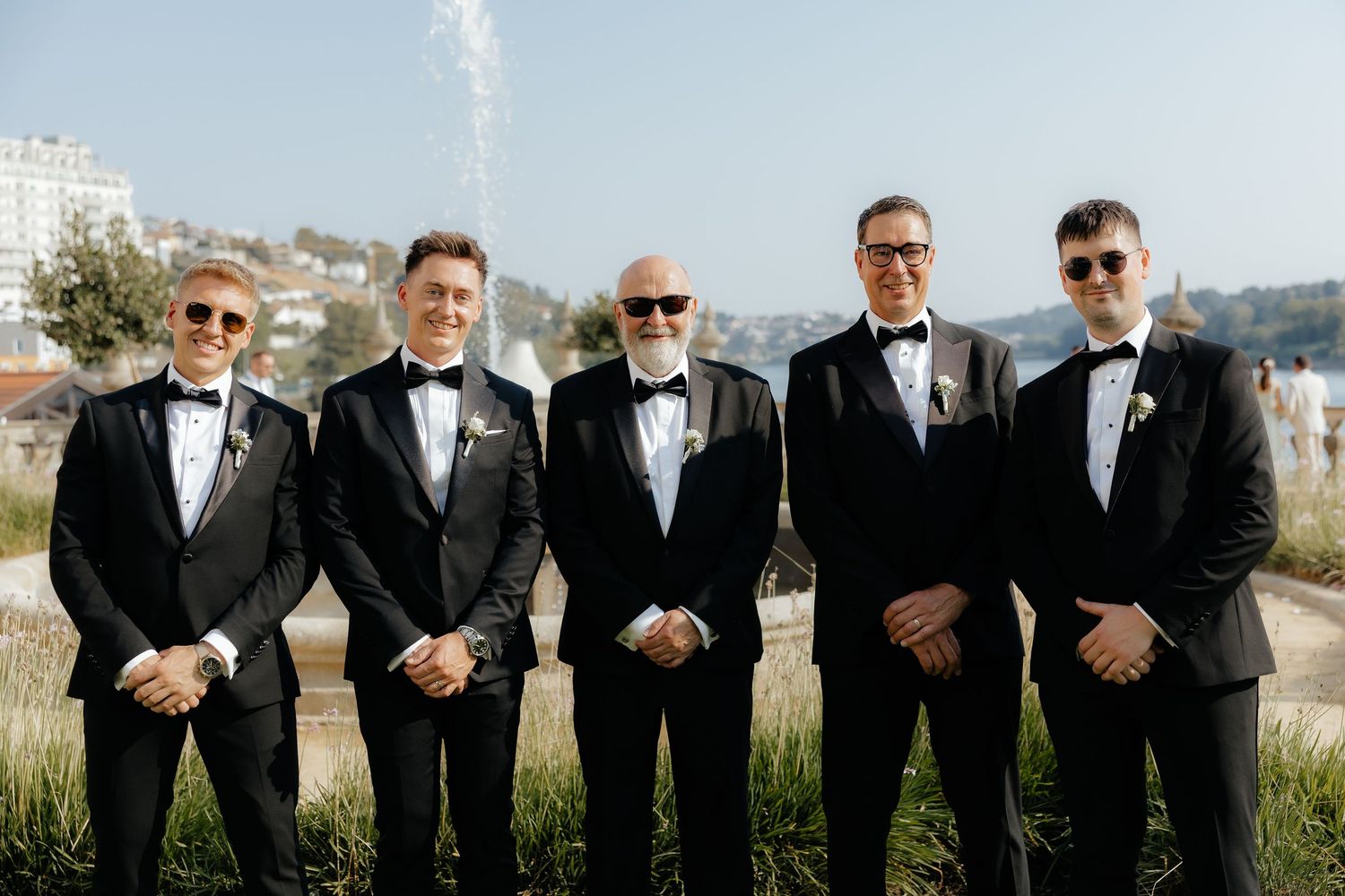 A group of groomsmen in black suits and sunglasses pose together outdoors in a fun candid style.