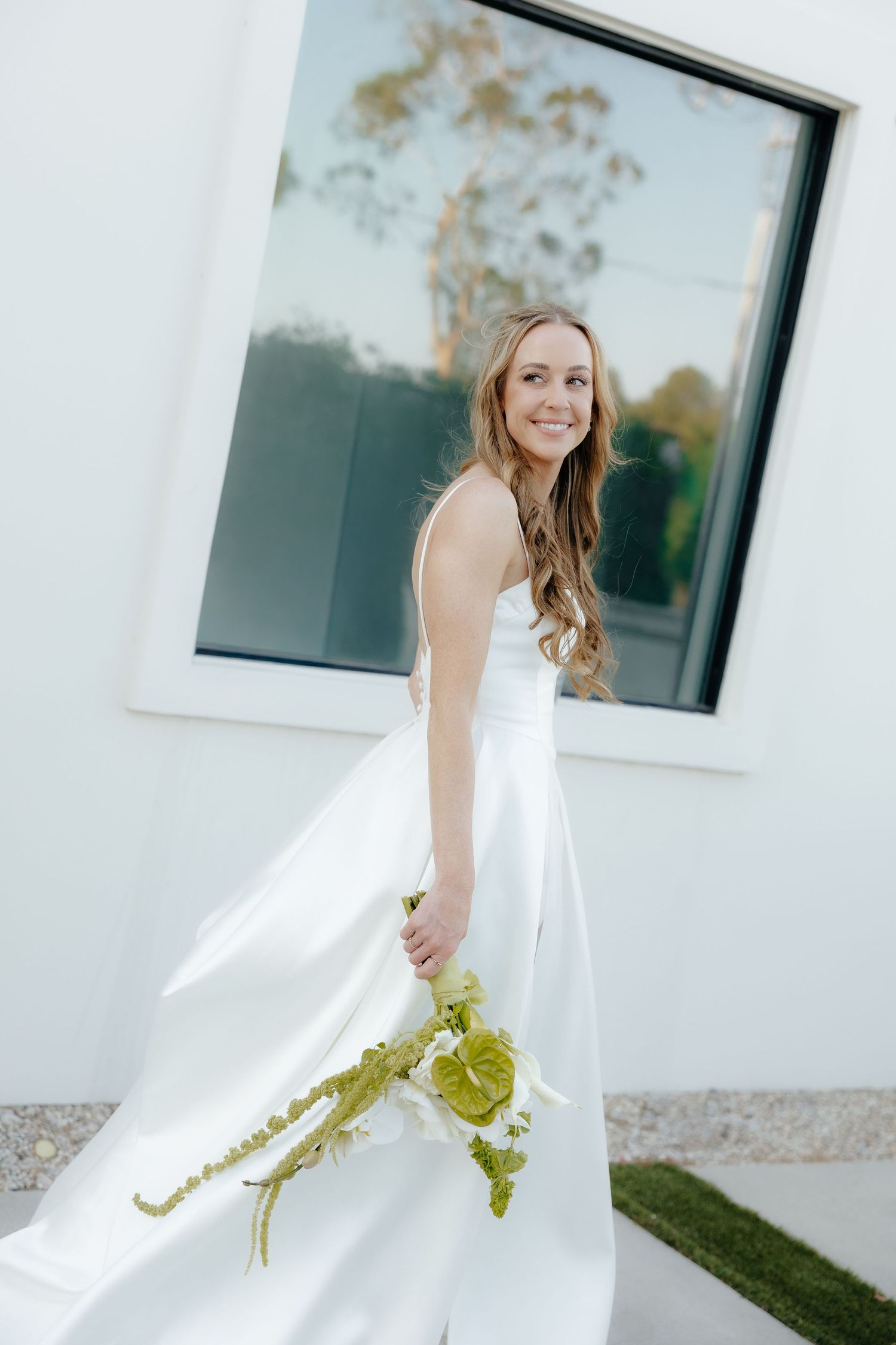 Smiling bride in white satin gown holding green bouquet posing before modern white building.