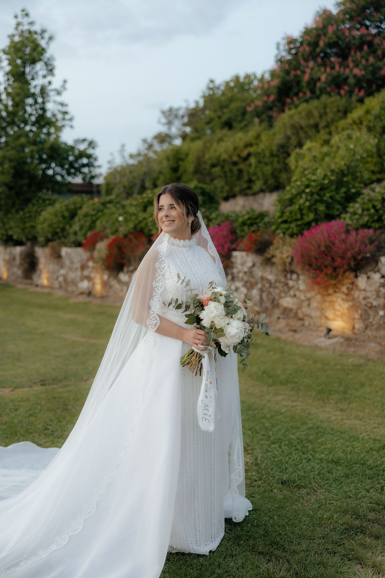Elegant bride in white beaded gown with veil holding floral bouquet in lush garden setting.
