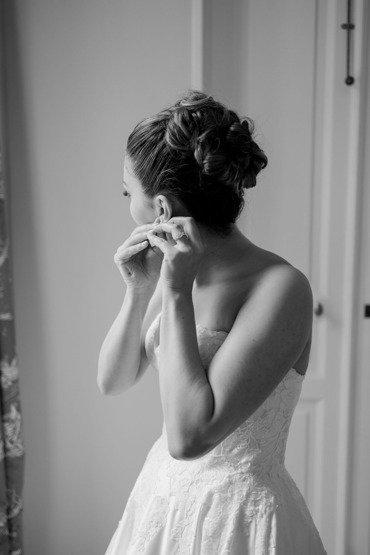 Black and white photo of a bride in a strapless lace gown putting on earrings, with an elegant updo hairstyle.