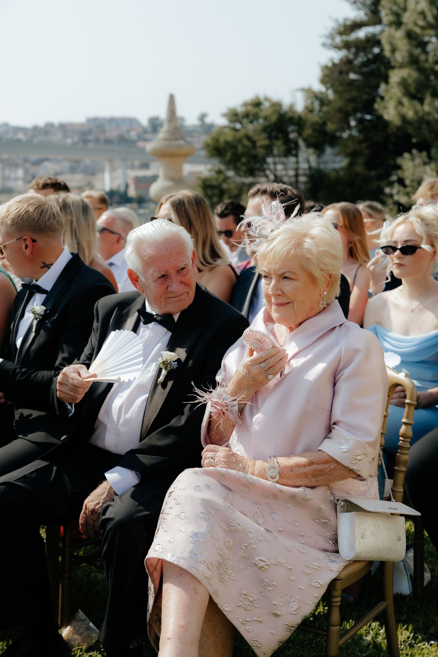 Elegant senior couple seated among wedding guests outdoors on a sunny ceremony day.