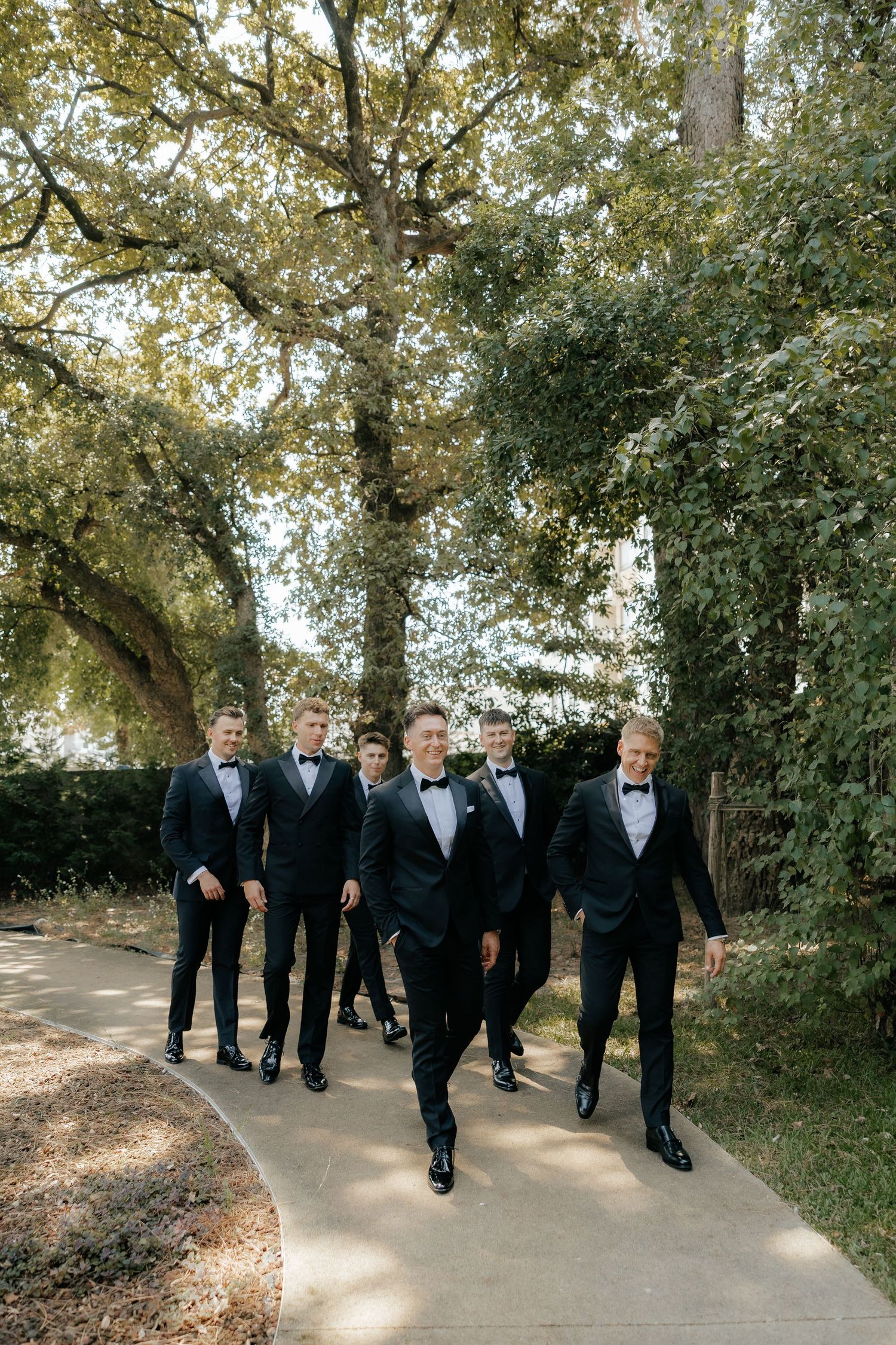 Six groomsmen in black tuxedos with bow ties walking confidently along a garden pathway surrounded by lush trees.