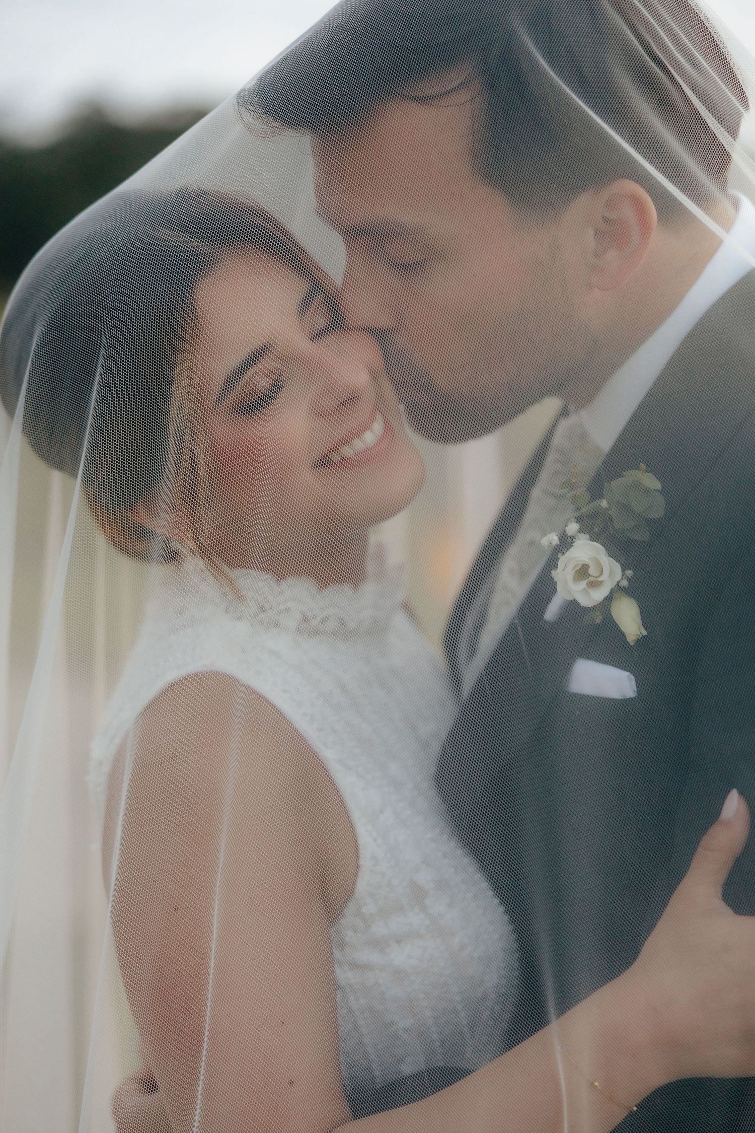 Bride smiling under veil as groom kisses her cheek in romantic wedding portrait.