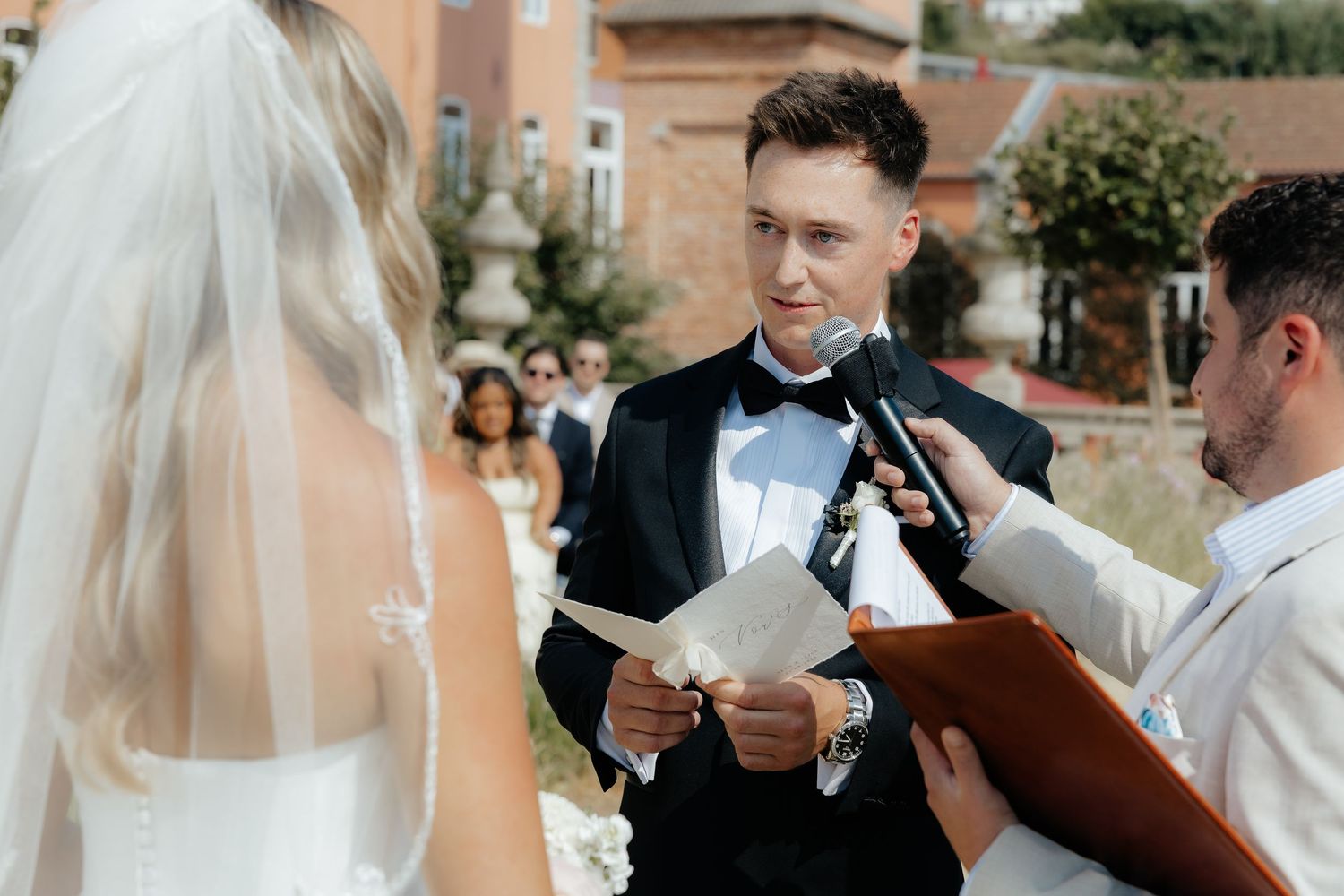 A group of wedding guests gather outdoors in formal attire while reading from papers during a daytime celebration.