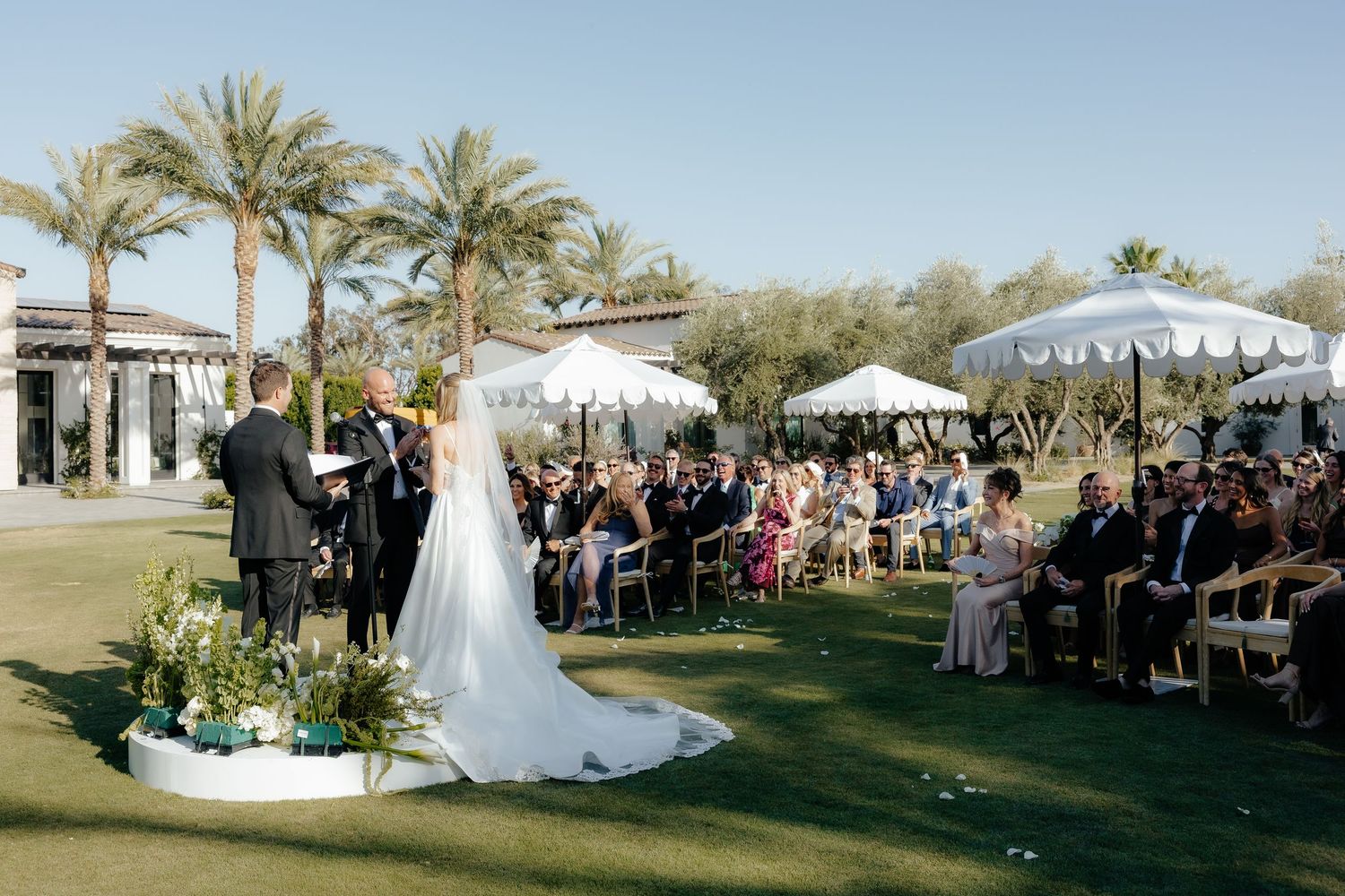 Bride and groom exchange vows on circular white platform at outdoor wedding ceremony surrounded by seated guests and palms.
