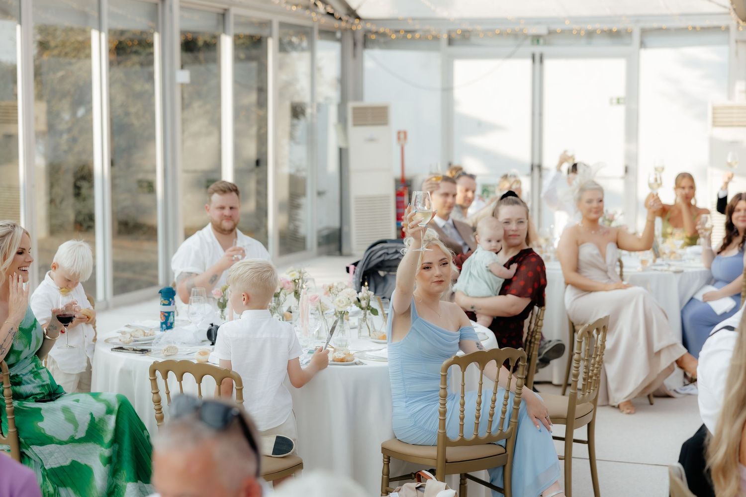 Wedding guests seated at elegant white-clothed tables in a bright, airy reception venue applaud during celebration.