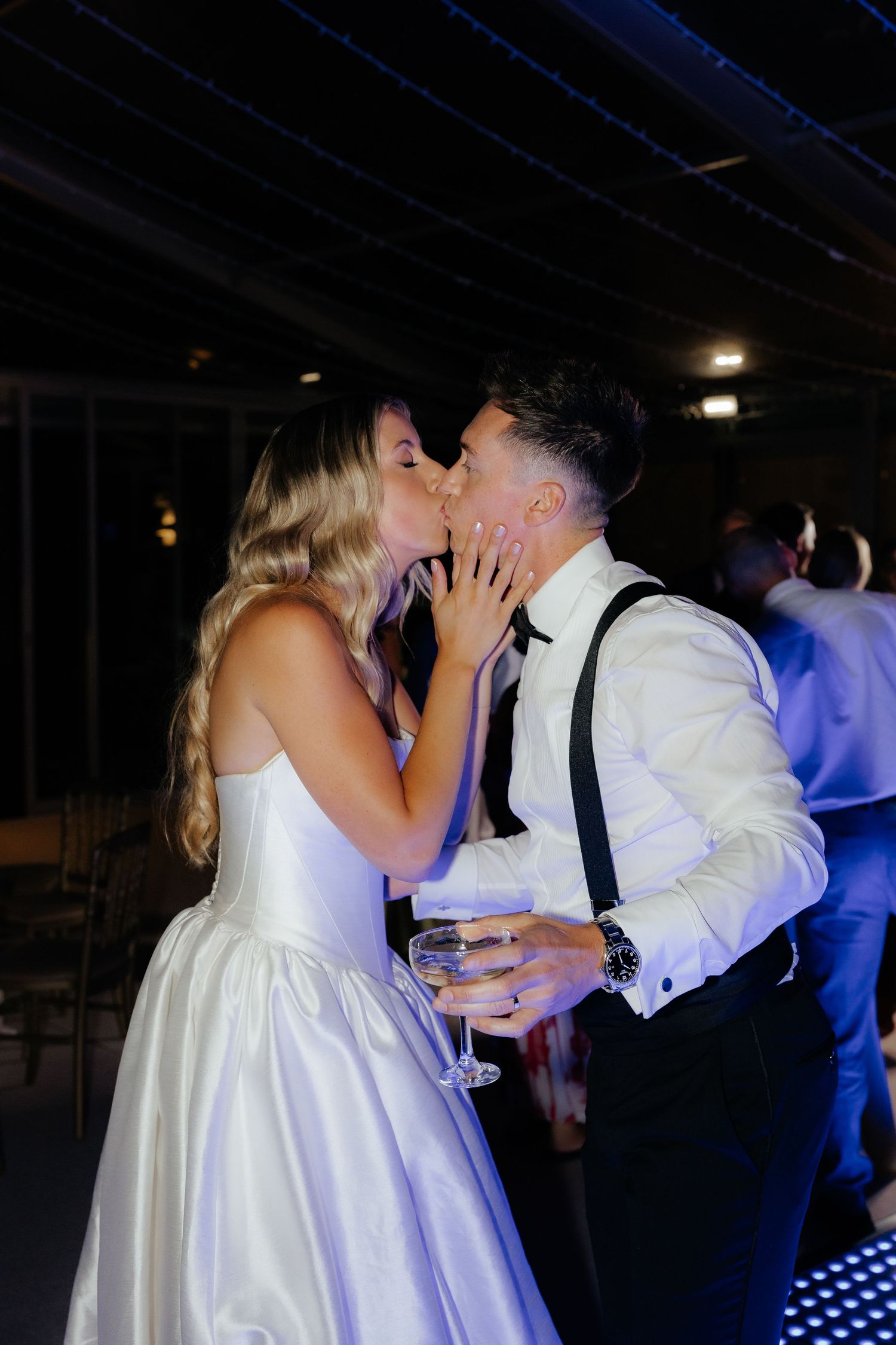 A couple shares a romantic kiss on their wedding day, with the bride in white and groom in suspenders holding a drink.