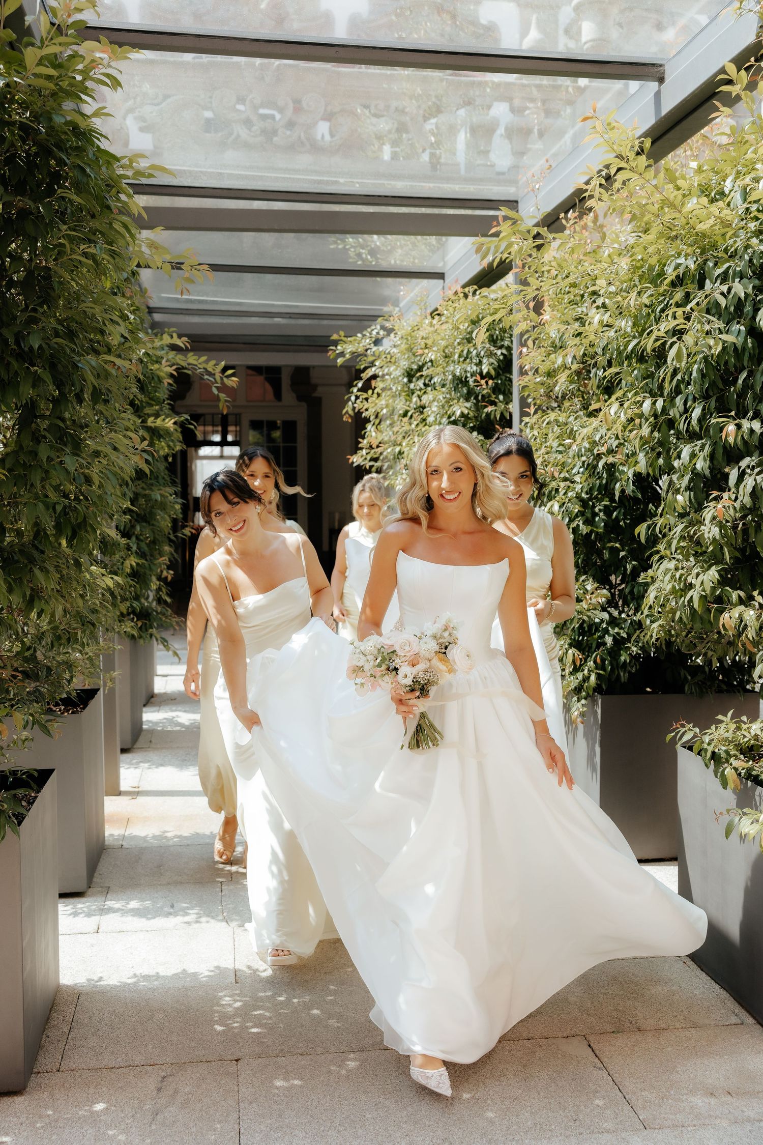 Bridesmaids in flowing white dresses walk through a covered walkway with lush greenery overhead on a sunny day.