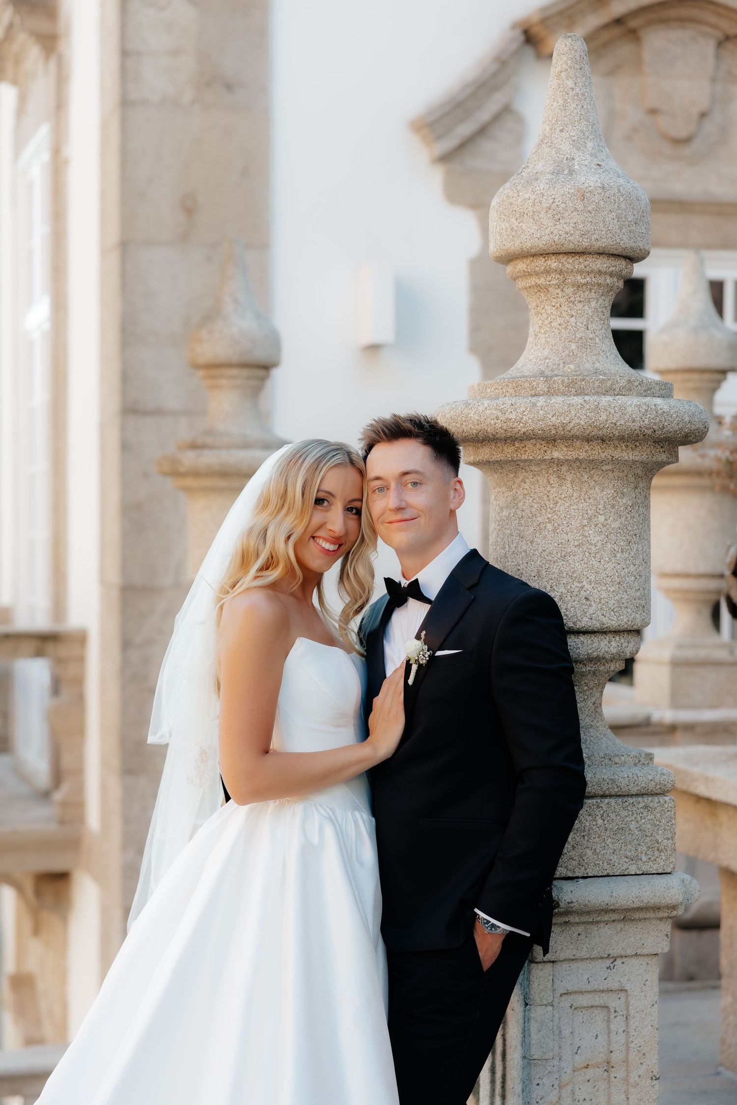 A bride in white and groom in black share romantic moments against stone pillars at an elegant venue.
