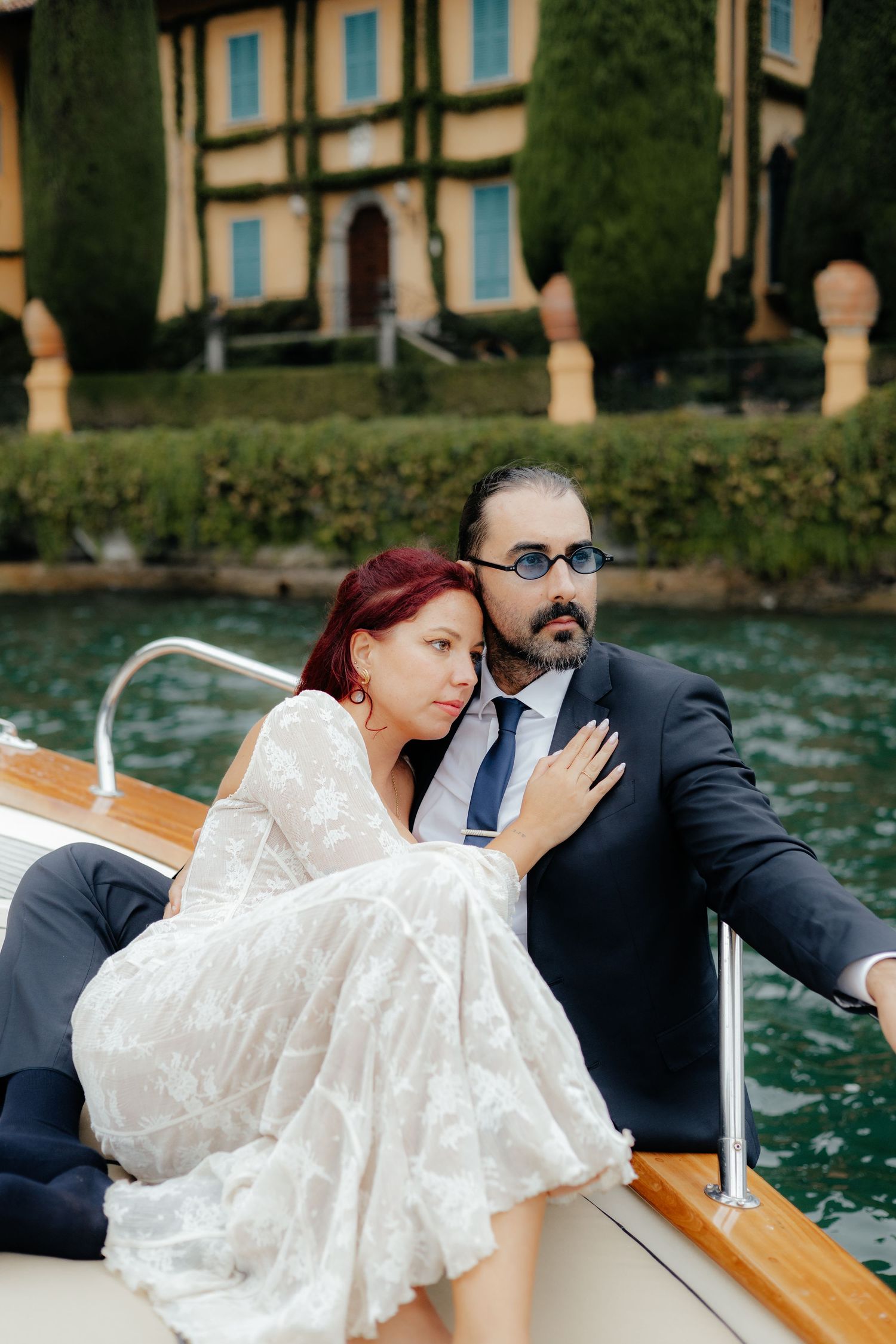 Bride in lace dress and groom in navy suit cuddle on a boat with Italian villa backdrop.