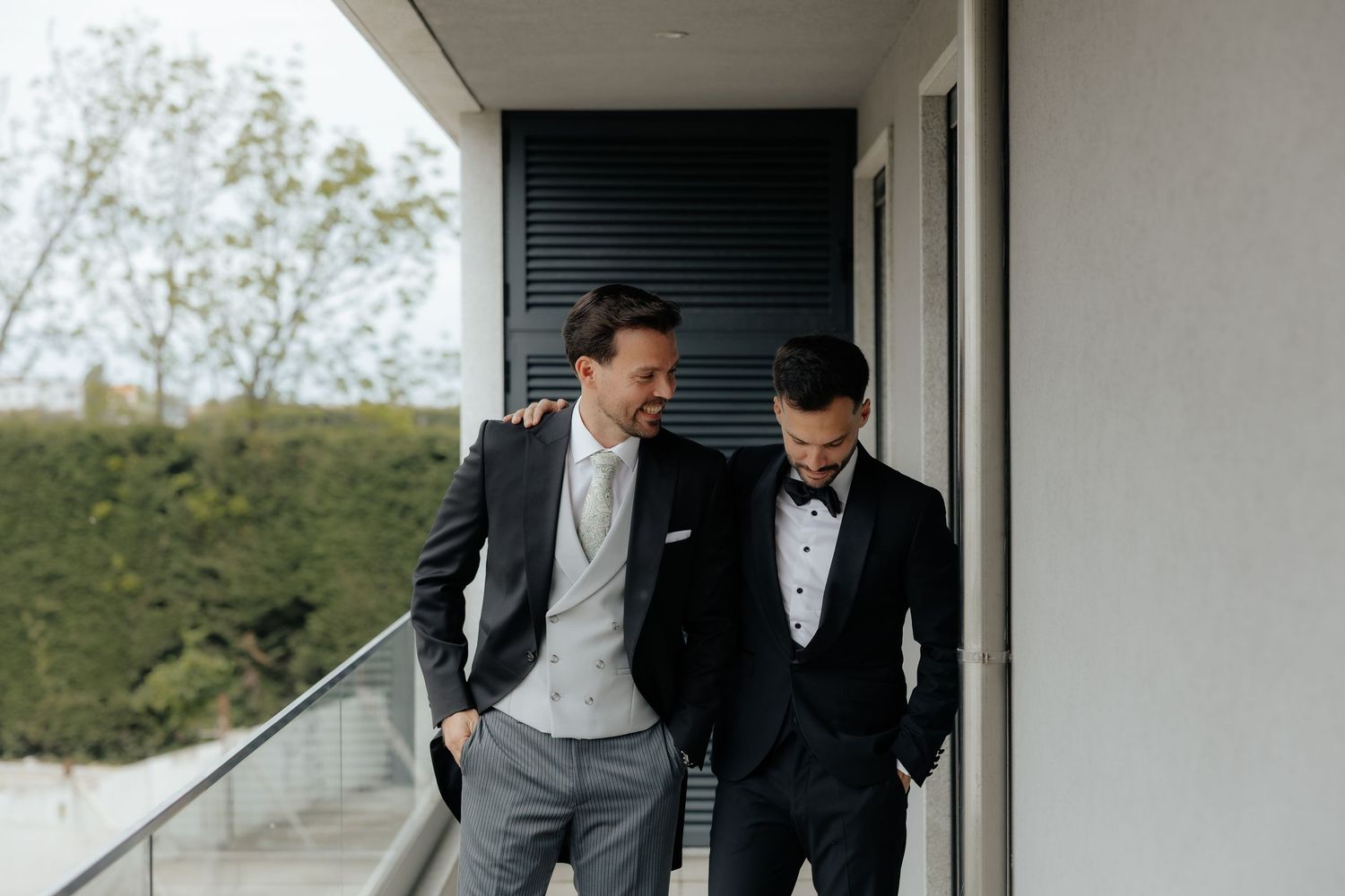 Two groomsmen in formal suits share a laugh on a modern balcony during wedding preparations.