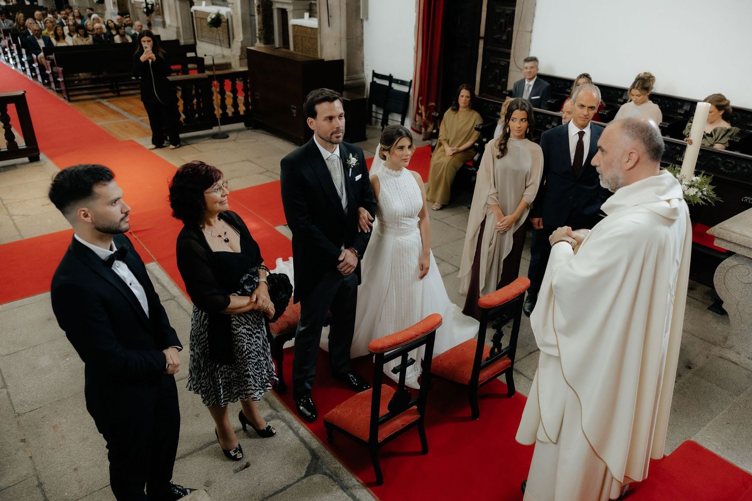 Wedding ceremony with bride, groom, and guests on red carpet before priest in church.