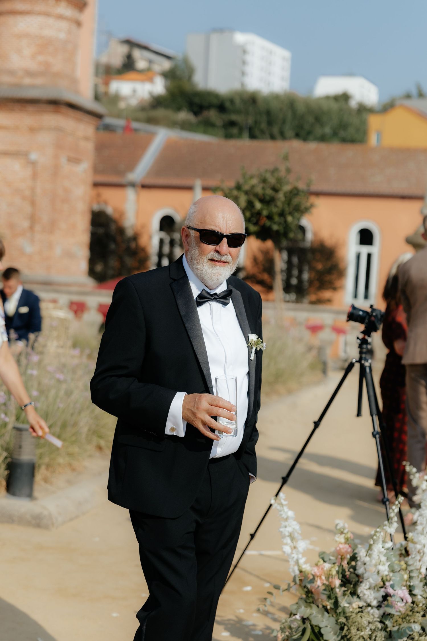 Distinguished older man in black tuxedo and sunglasses holds champagne glass at outdoor wedding.