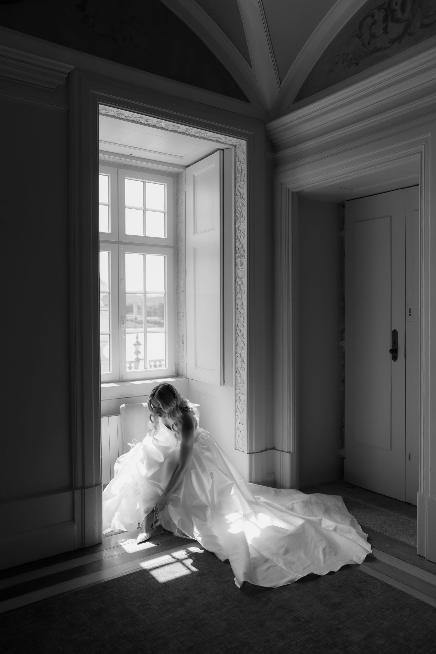Black and white artistic photo of bride in white gown sitting by ornate window in historic venue.