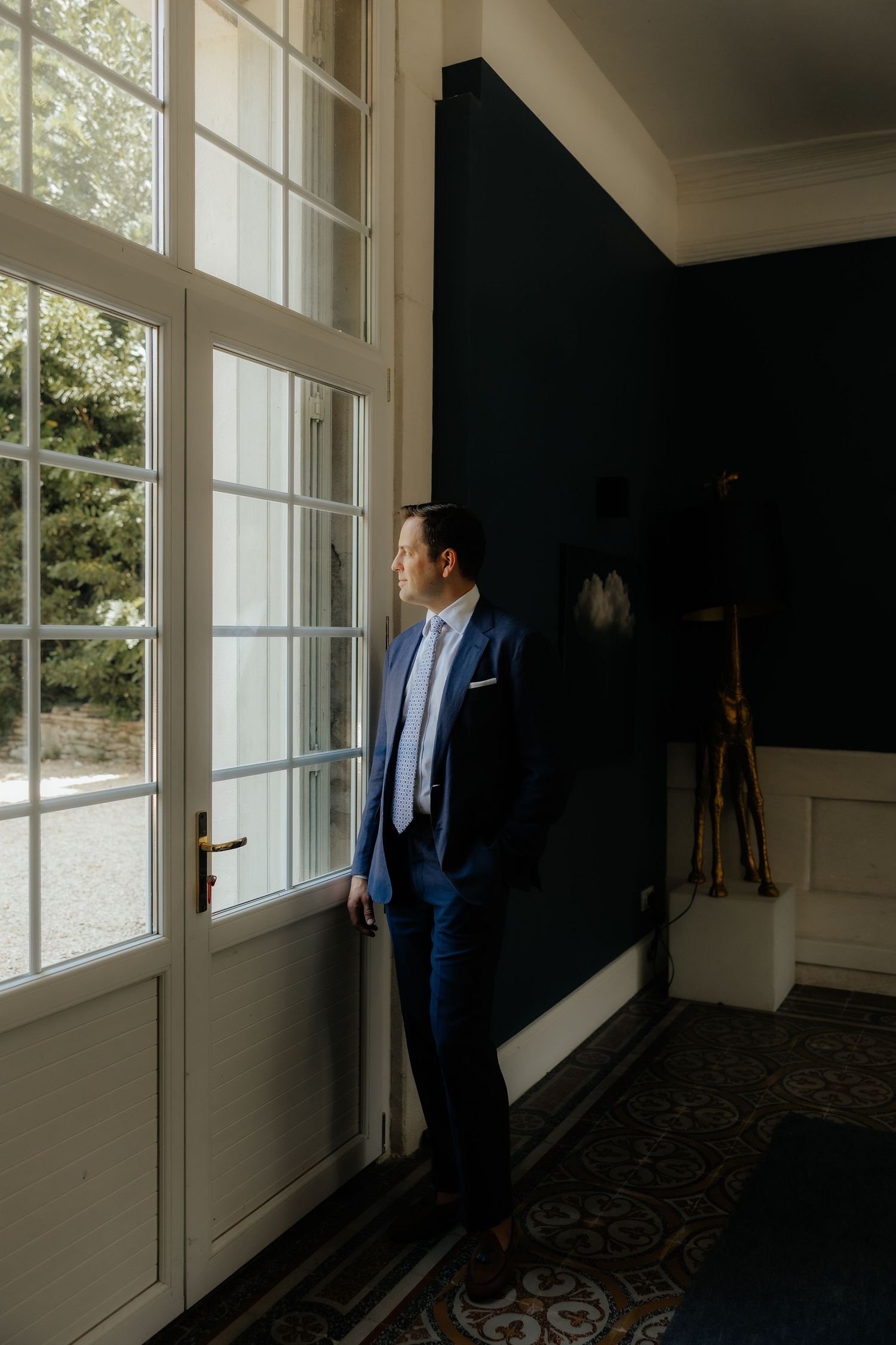 A person in a navy suit looks out large windows in an elegant room with natural lighting.