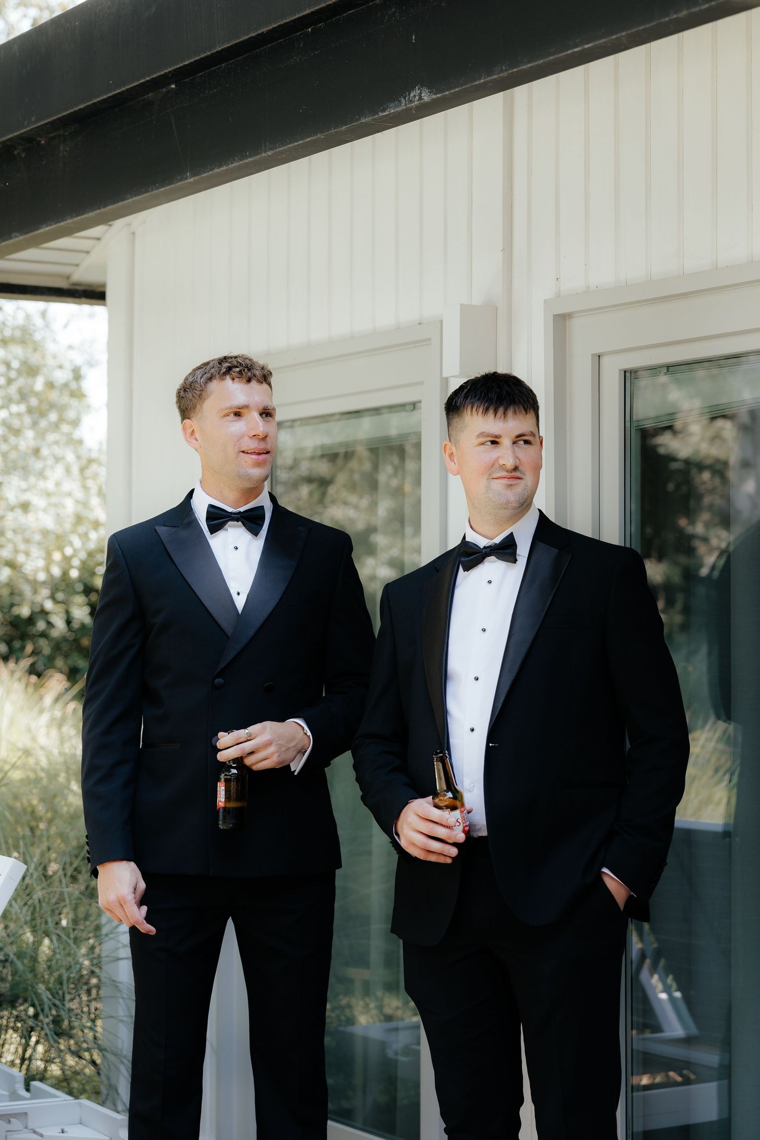 Two people in black tuxedos share a laugh while holding drinks at a formal event.