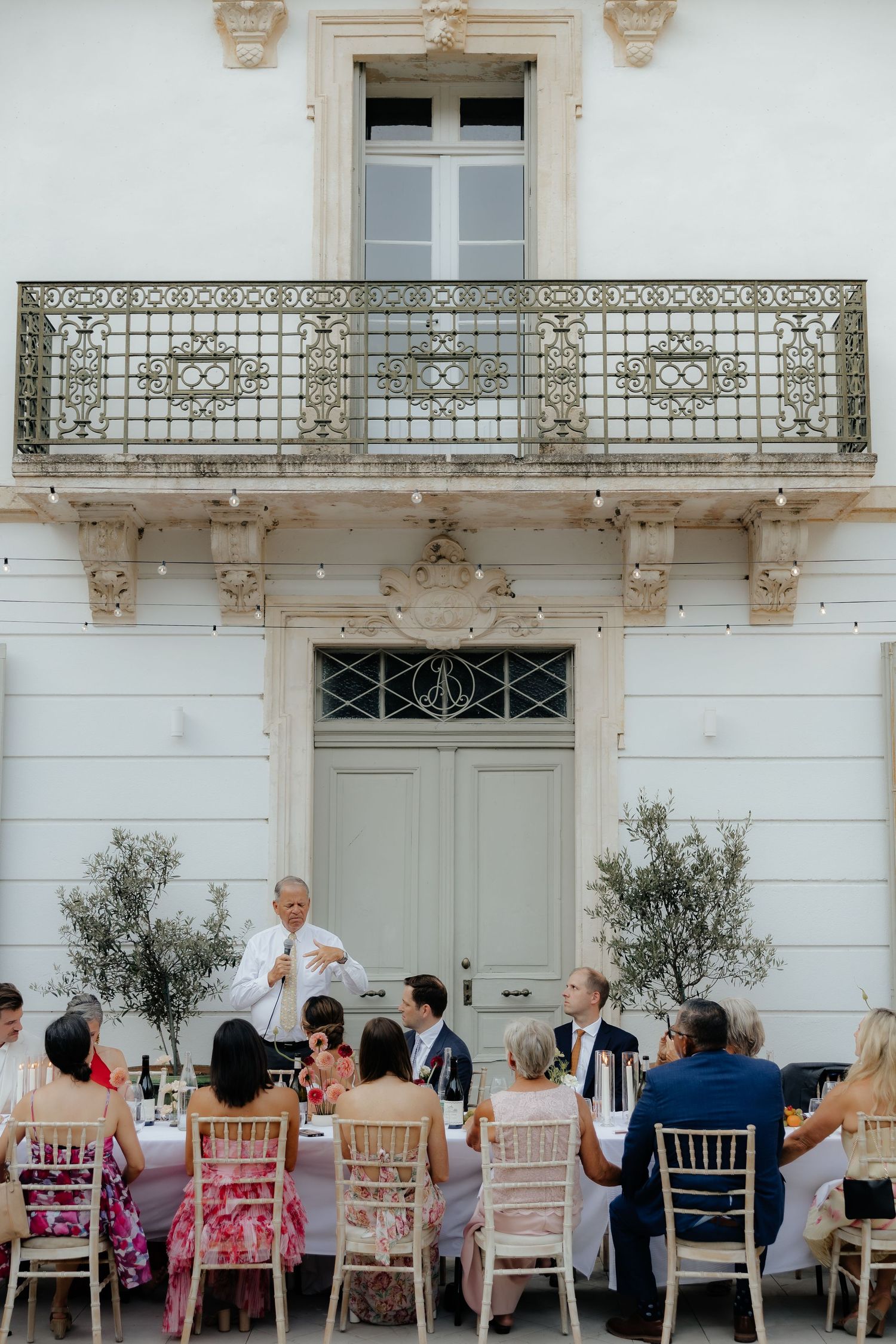 Wedding guests seated at an elegant outdoor reception with white chairs and olive trees against a classical building facade.