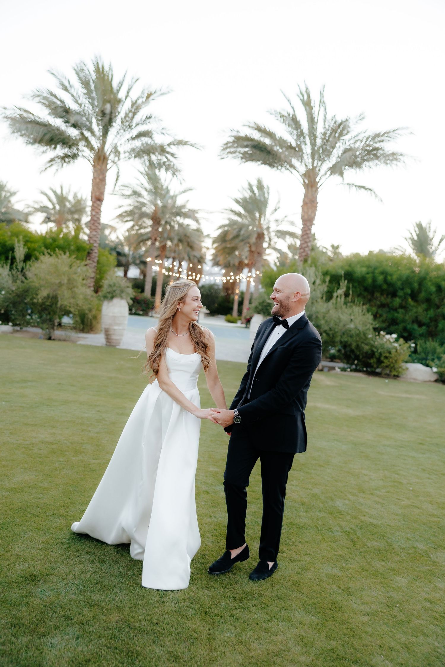 Bride and groom holding hands facing each other on lush lawn with palm trees and string lights behind.