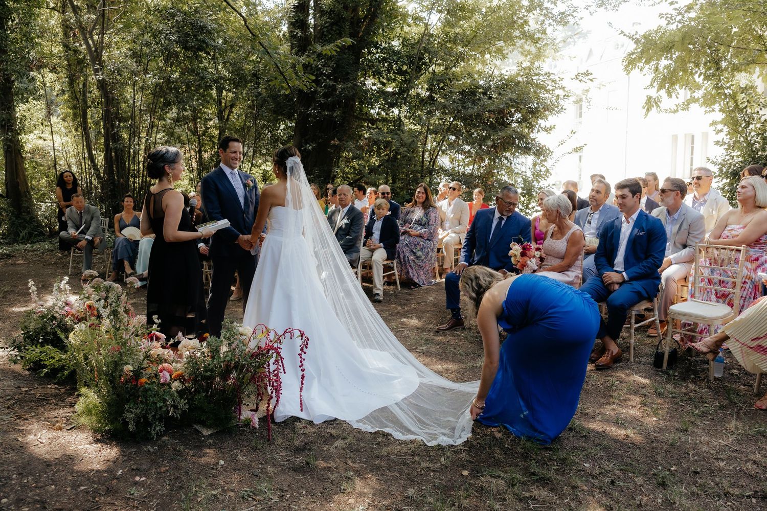 An outdoor wedding ceremony with guests seated on white chairs under trees.