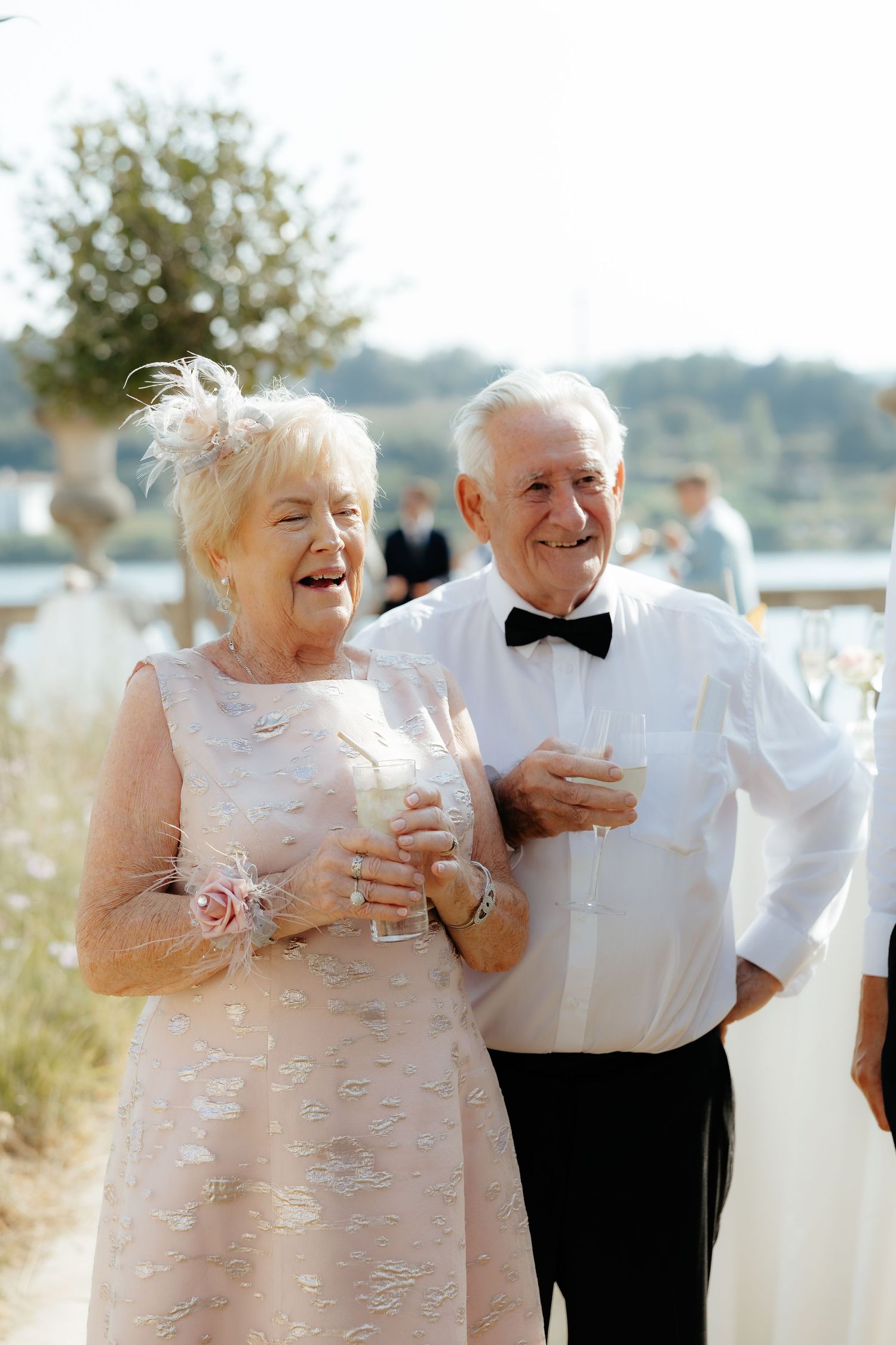 Joyful elderly couple in formal wedding attire smile and hold drinks at outdoor wedding reception.