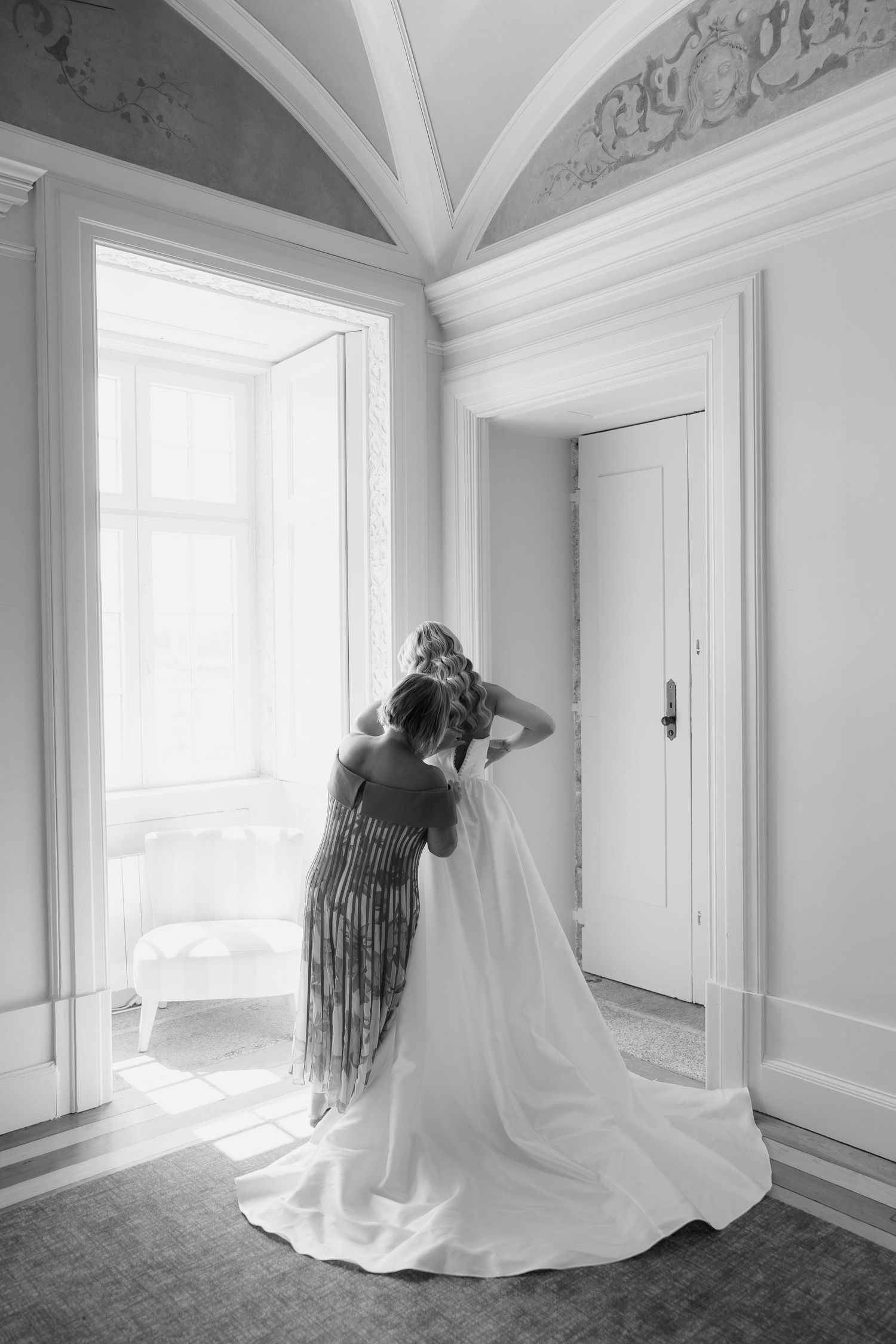 Black and white photo of bride getting veil adjusted by bridesmaid in elegant room with arched ceiling.