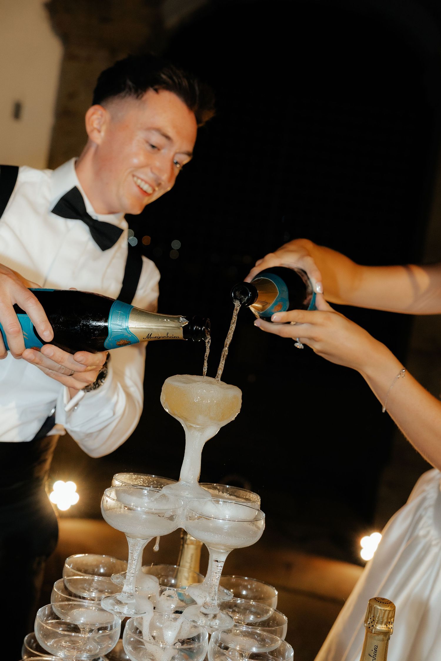 Smiling groom pours champagne tower at wedding reception with bride helping fill stacked coupe glasses.