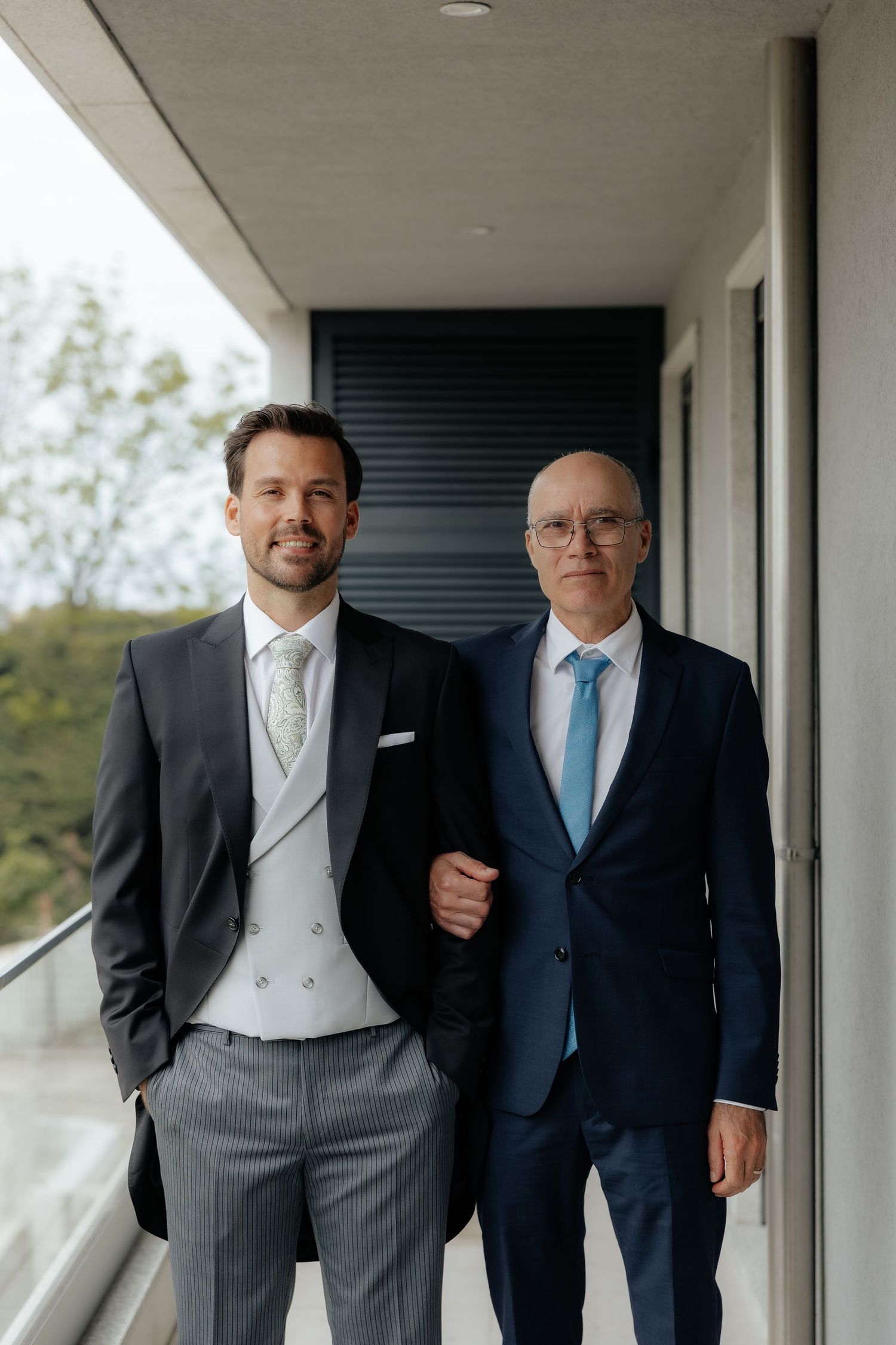Groom in black tuxedo poses with older man in navy suit on balcony.