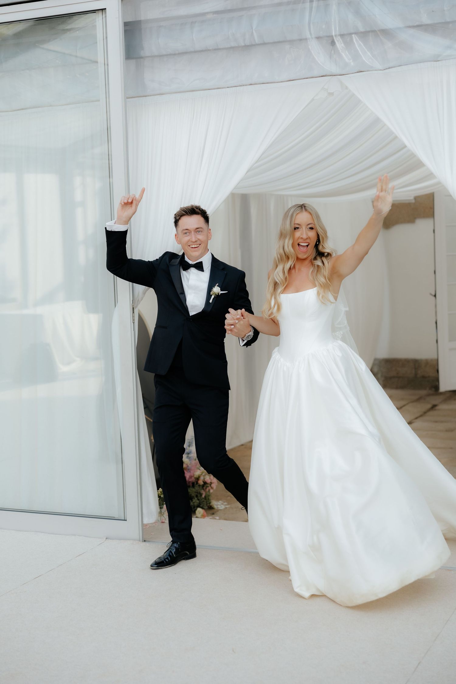 A joyful couple in wedding attire playfully poses against white curtains in a bright indoor setting.