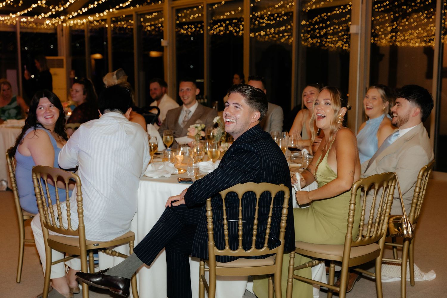 Wedding reception guests seated at long tables under warm string lights in an elegant indoor venue.