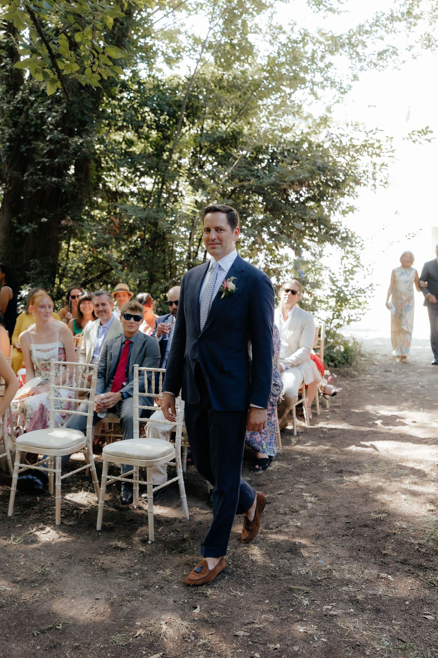 An outdoor wedding ceremony setup with white chairs along a tree-lined path in dappled sunlight.