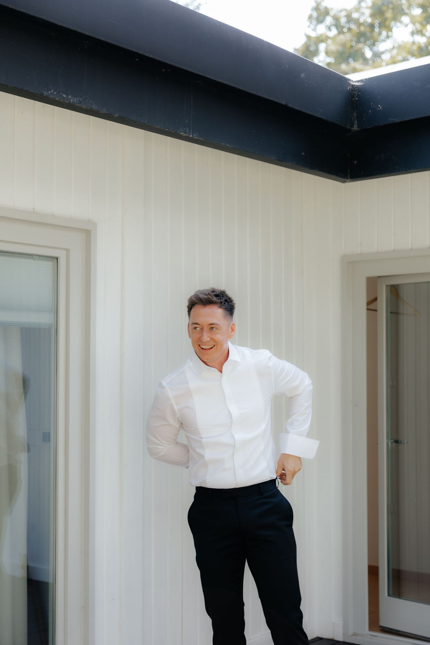 Person in white dress shirt and black pants poses playfully against a white wall in an interior space.