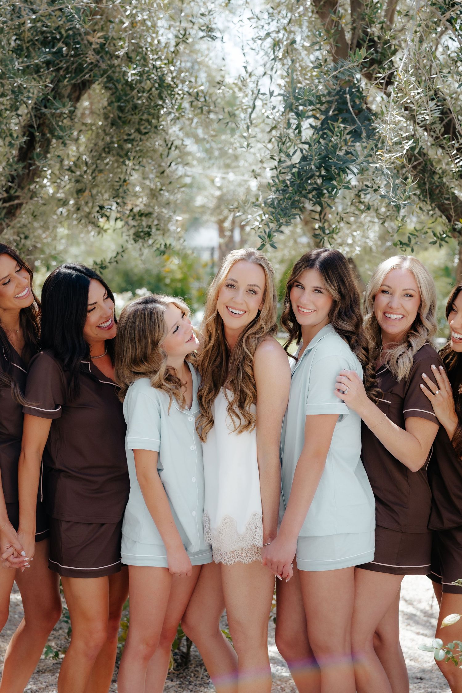 Bride in white lace romper surrounded by bridesmaids in brown and mint pajama sets under olive trees.