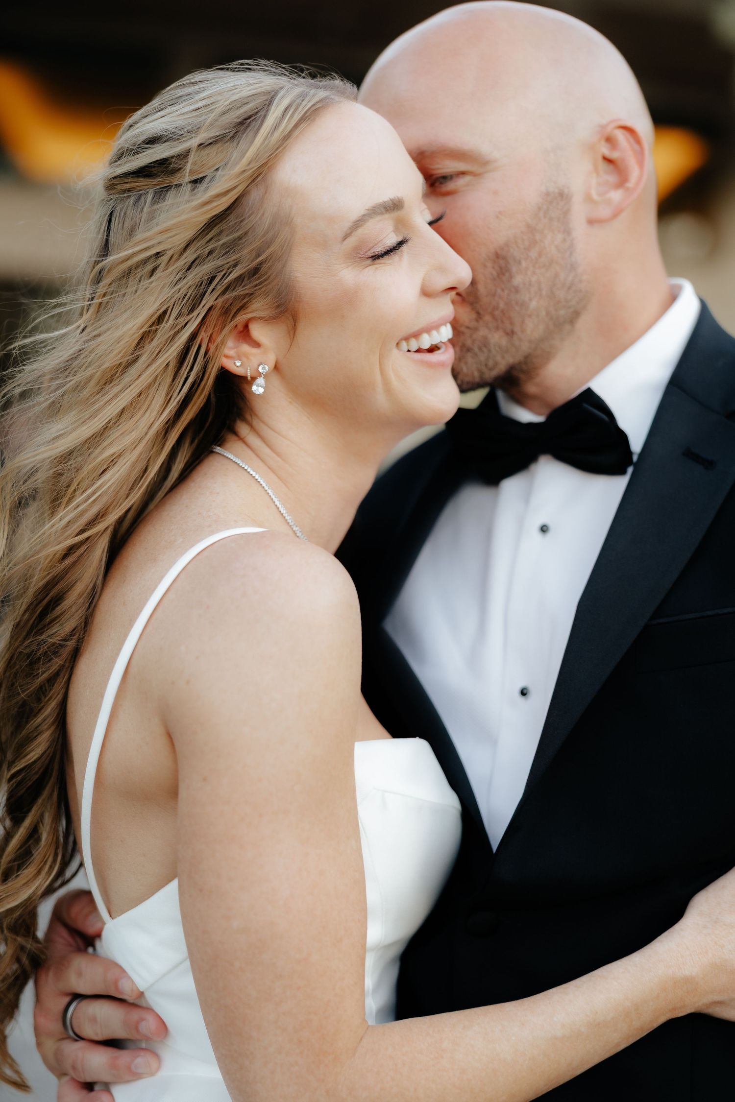 Close-up of groom kissing smiling bride on cheek, bride wearing pearl earrings and white wedding gown.