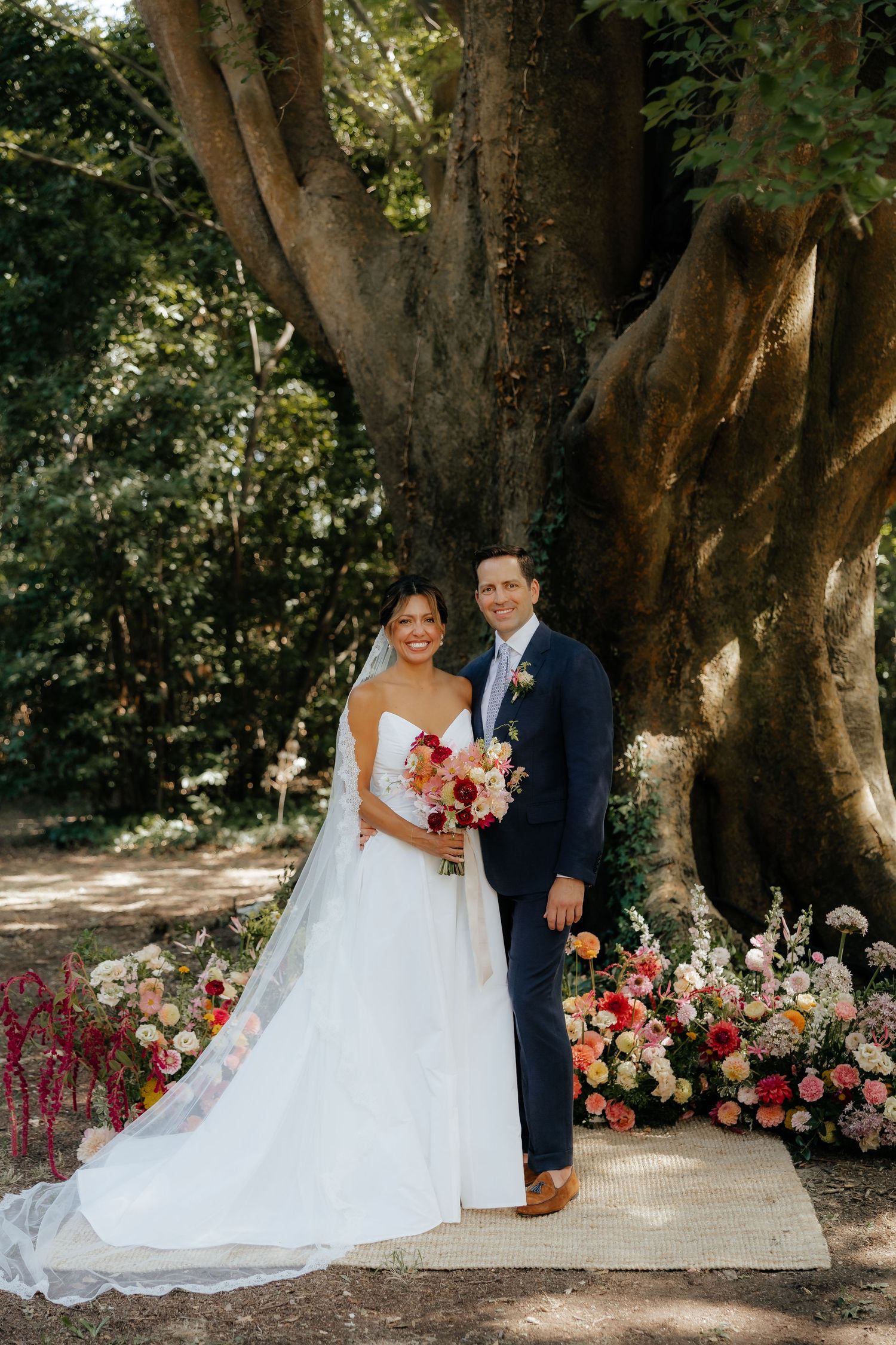 A wedding couple poses in front of a majestic tree surrounded by vibrant floral arrangements.