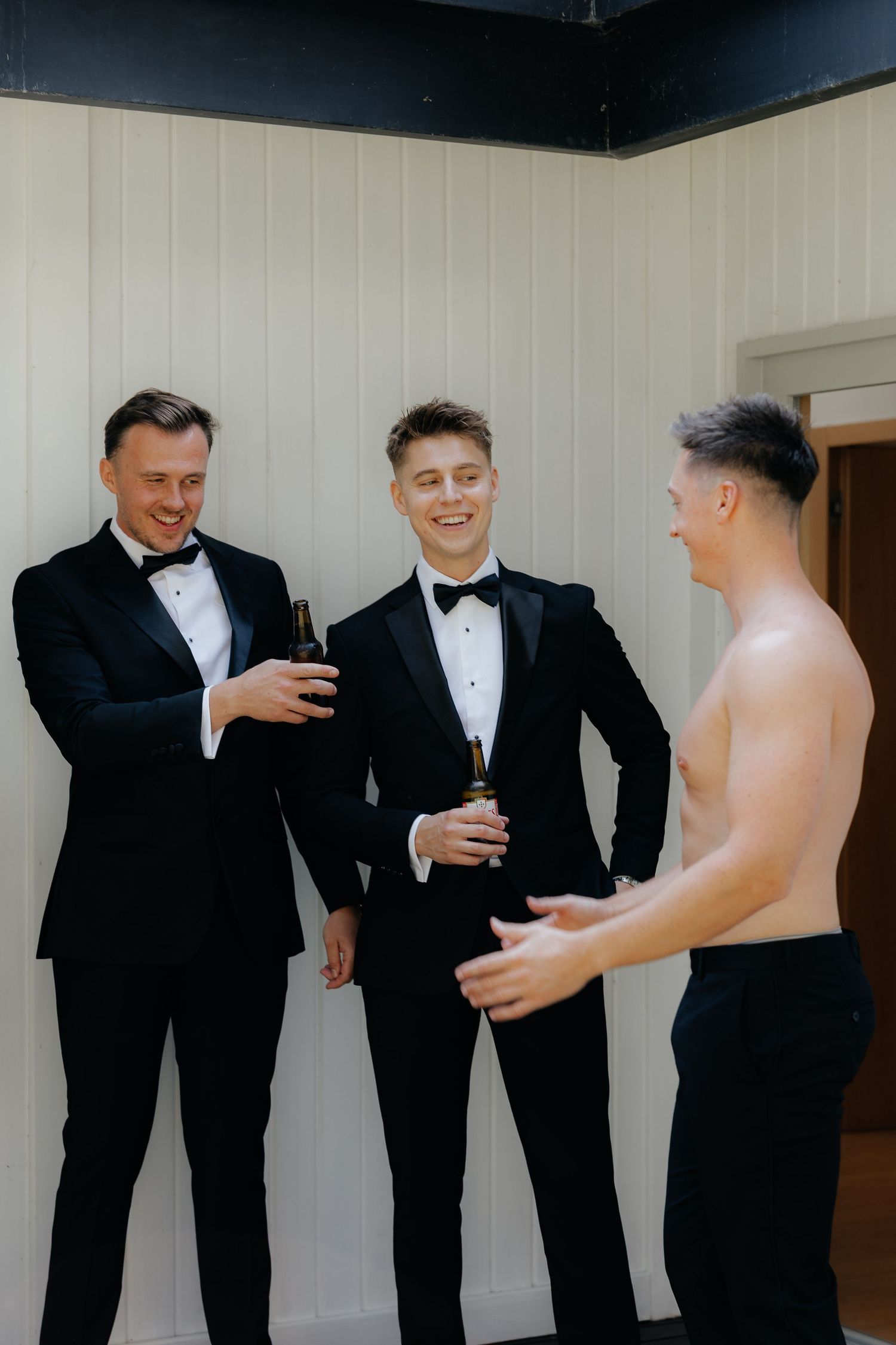 Three people in formal black tuxedos sharing drinks and laughing together during a celebration.