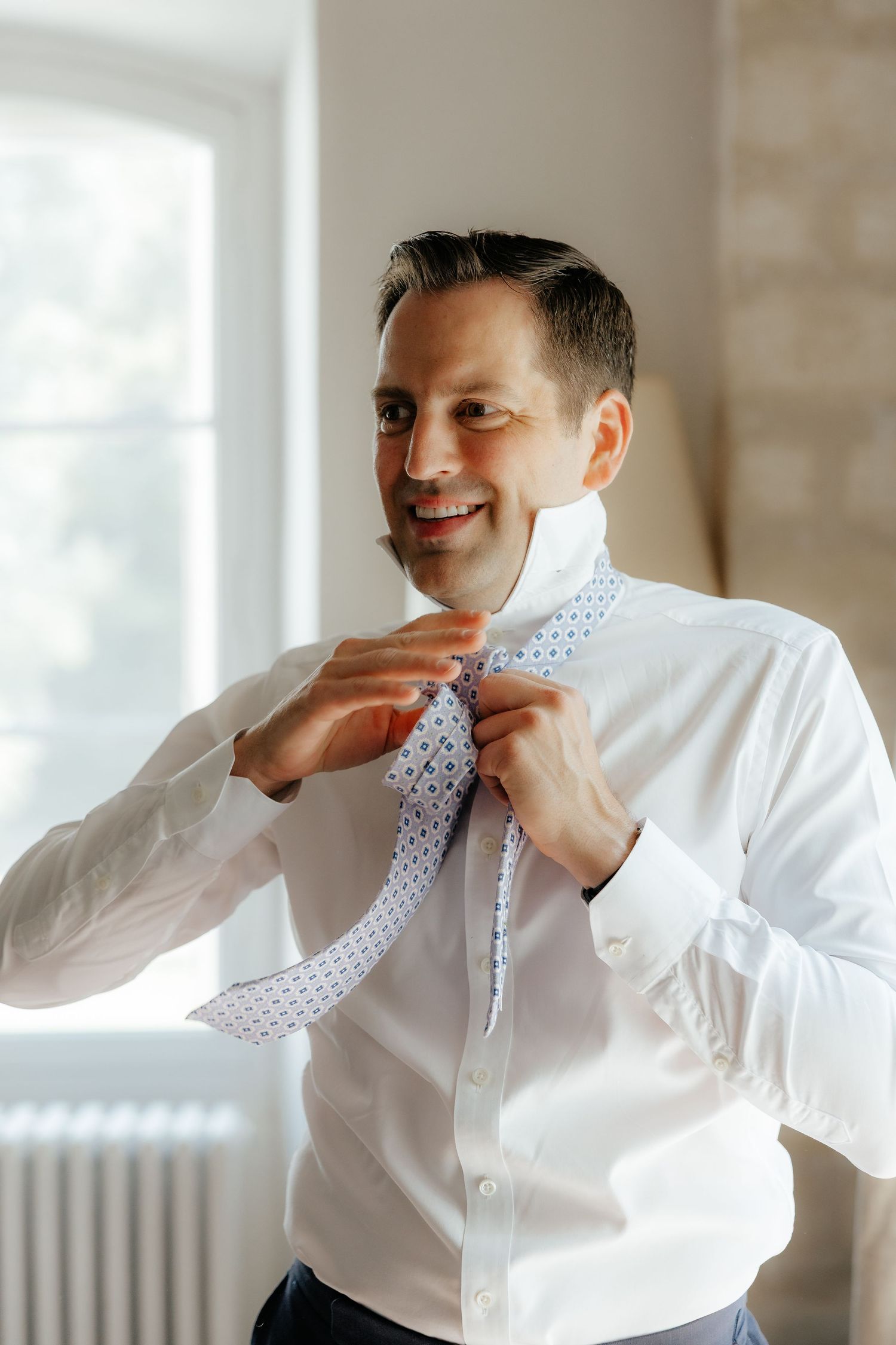 A person in a crisp white dress shirt adjusts a polka dot tie while getting ready near a window.