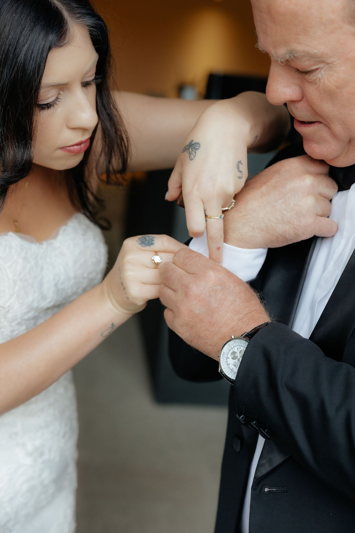 Bride with tattoos helping older man in tuxedo fasten cufflinks during wedding preparation.