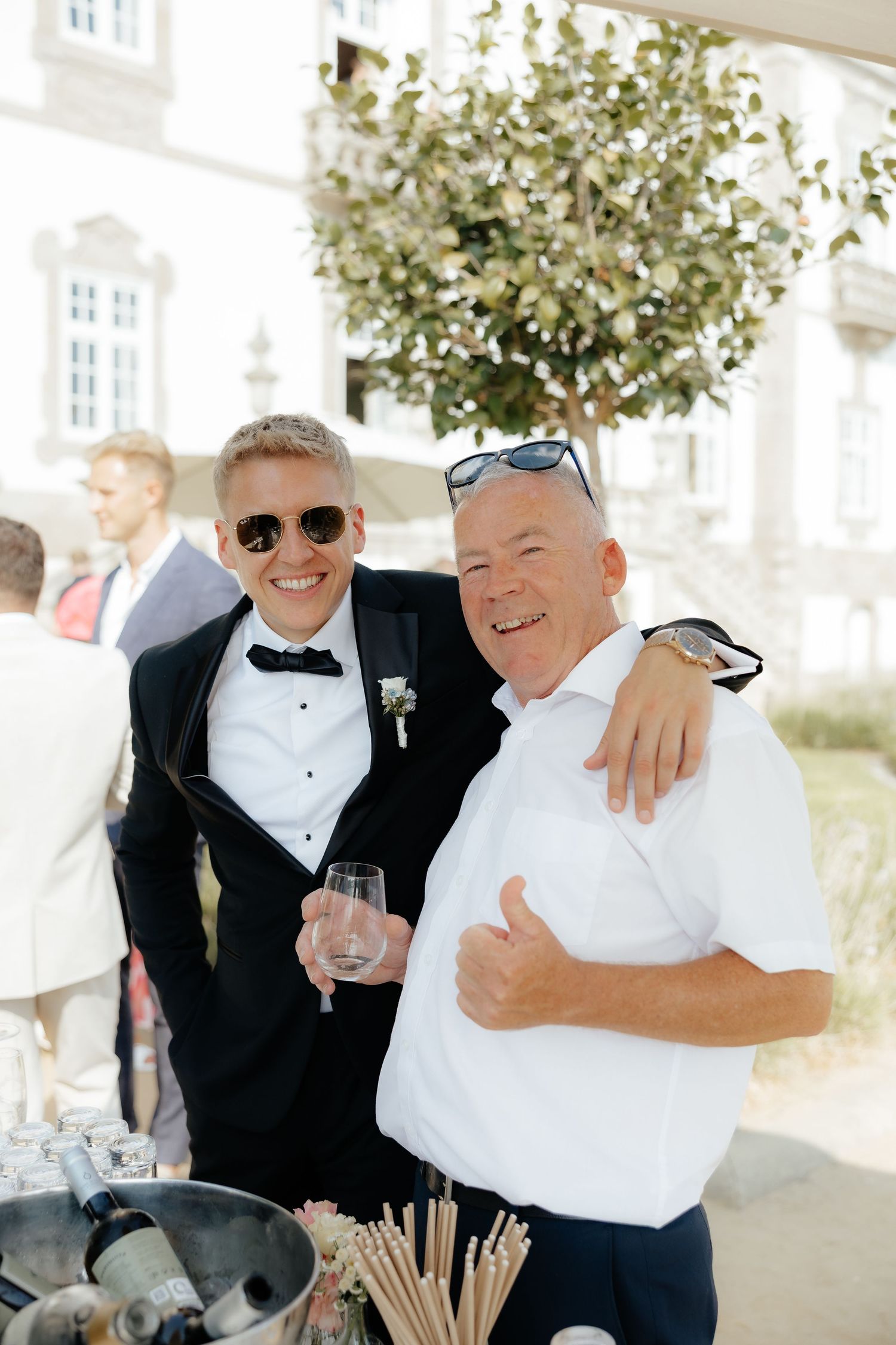 Groom in black tuxedo with sunglasses poses with older man in white shirt at outdoor wedding.
