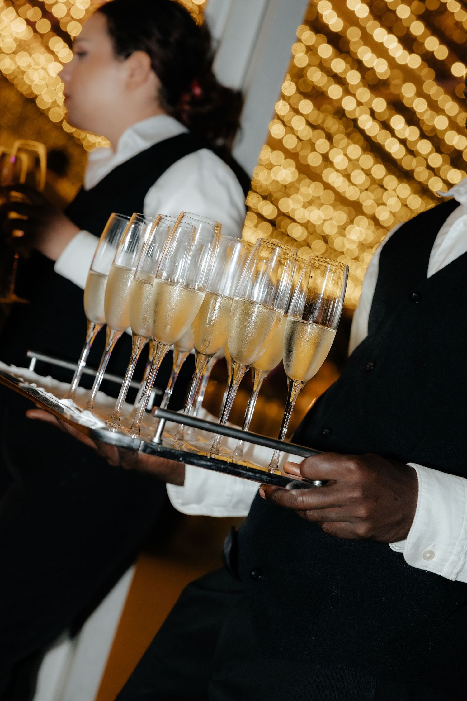 Wedding server carrying a silver tray of champagne flutes with golden bokeh lights in the background.