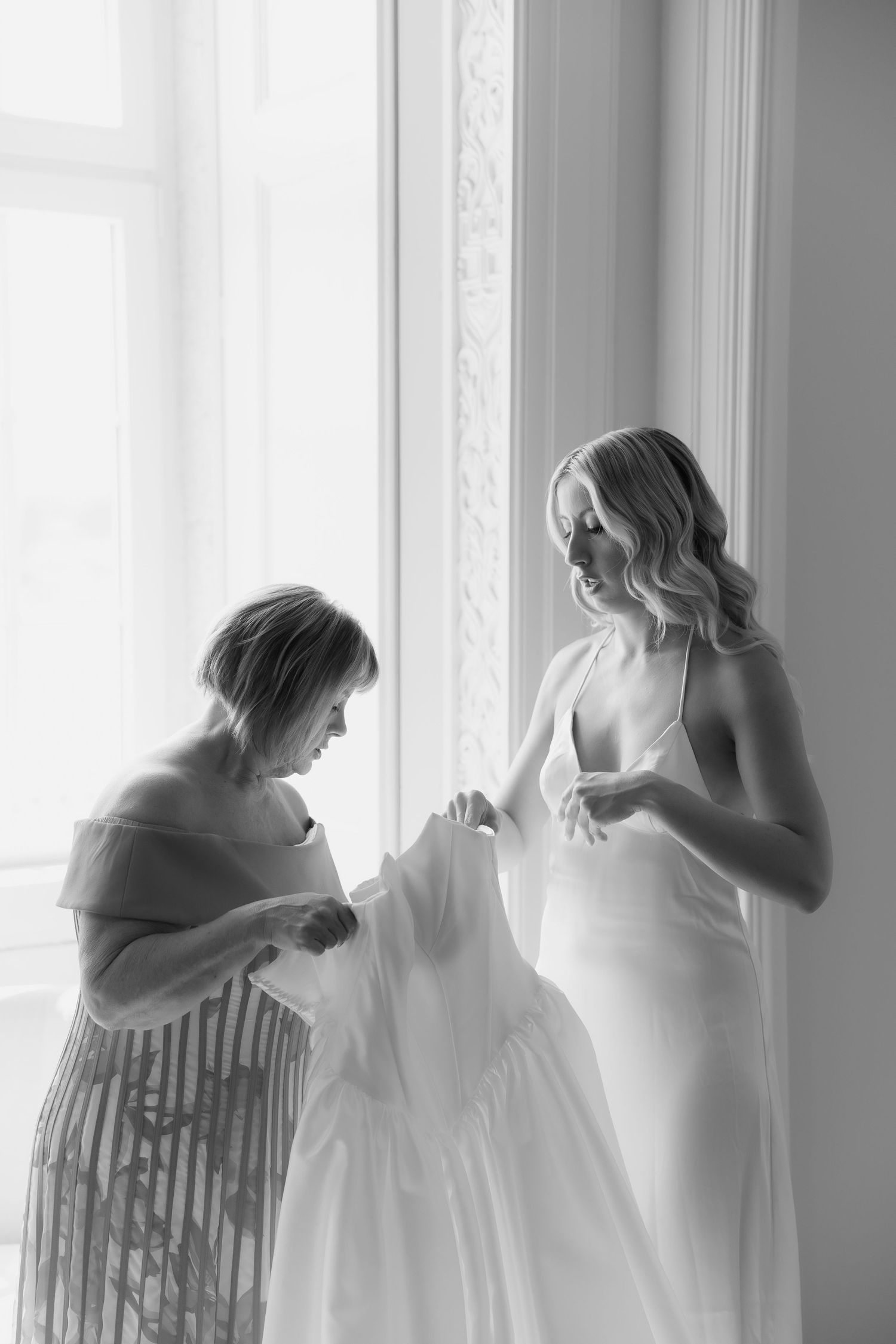Black and white photo of mother helping smiling bride with her flowing white wedding dress.