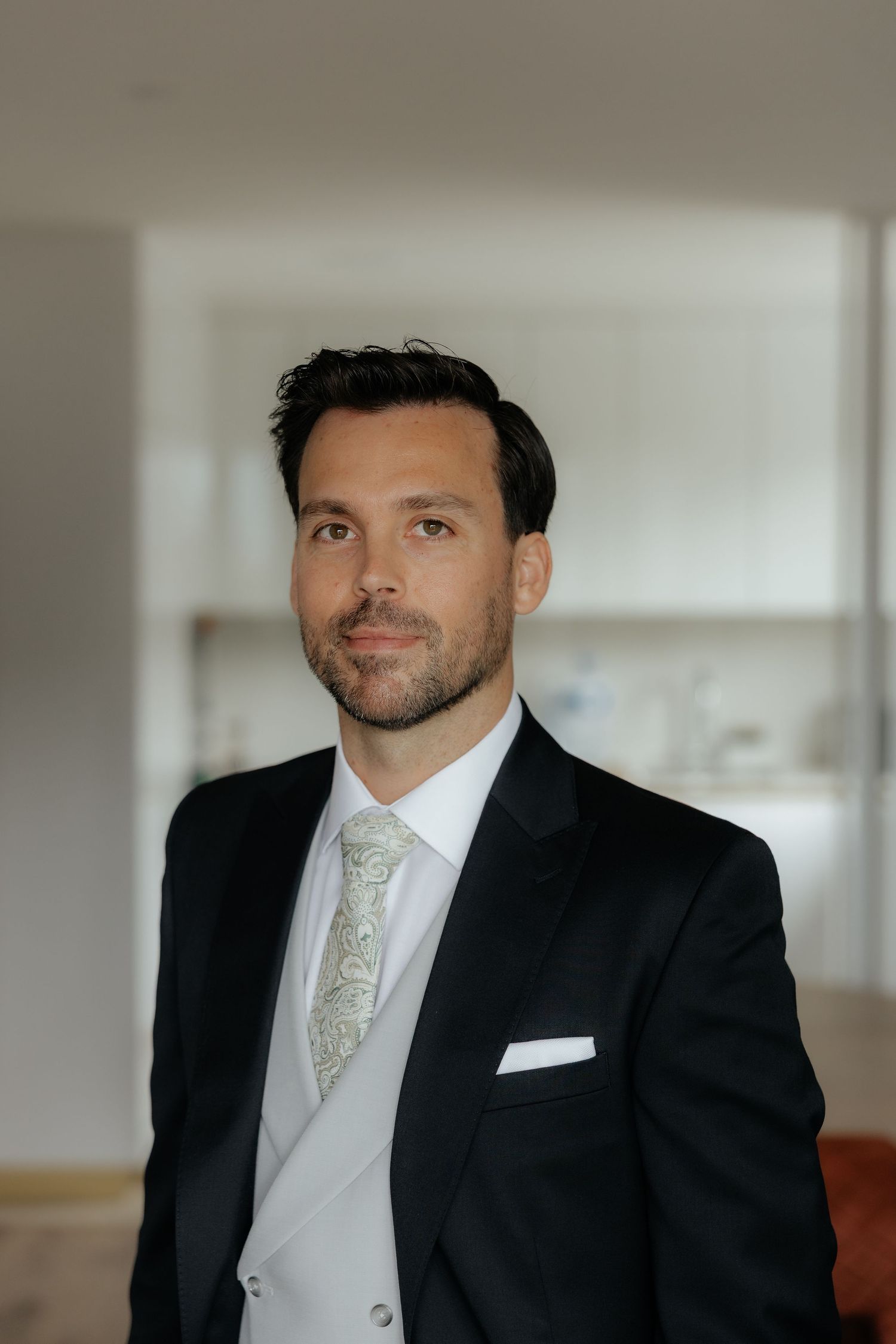 Groom in black tuxedo with white vest and paisley tie poses indoors.