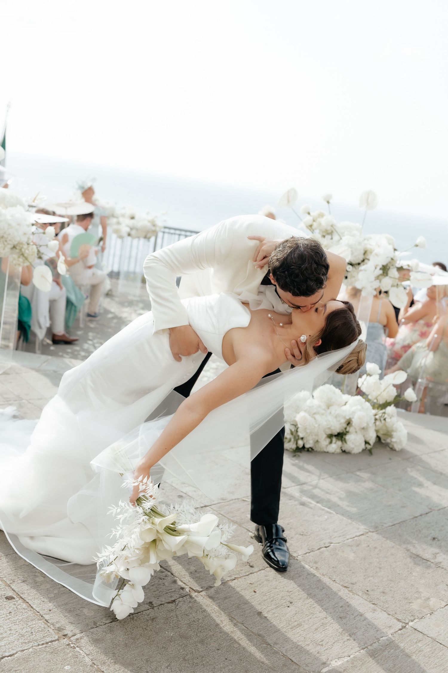 Groom dips bride for a romantic kiss at outdoor wedding ceremony with white floral arrangements and guests in background.