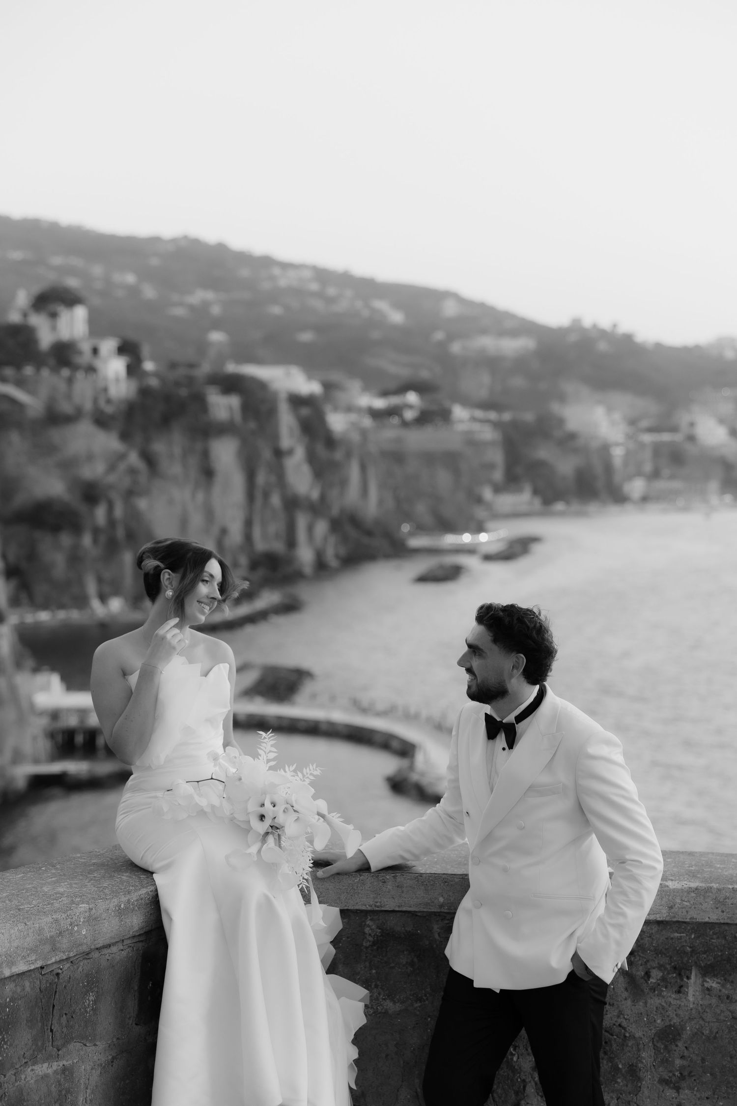 Black and white wedding photo of bride and groom on a cliffside terrace overlooking a scenic coastal bay.