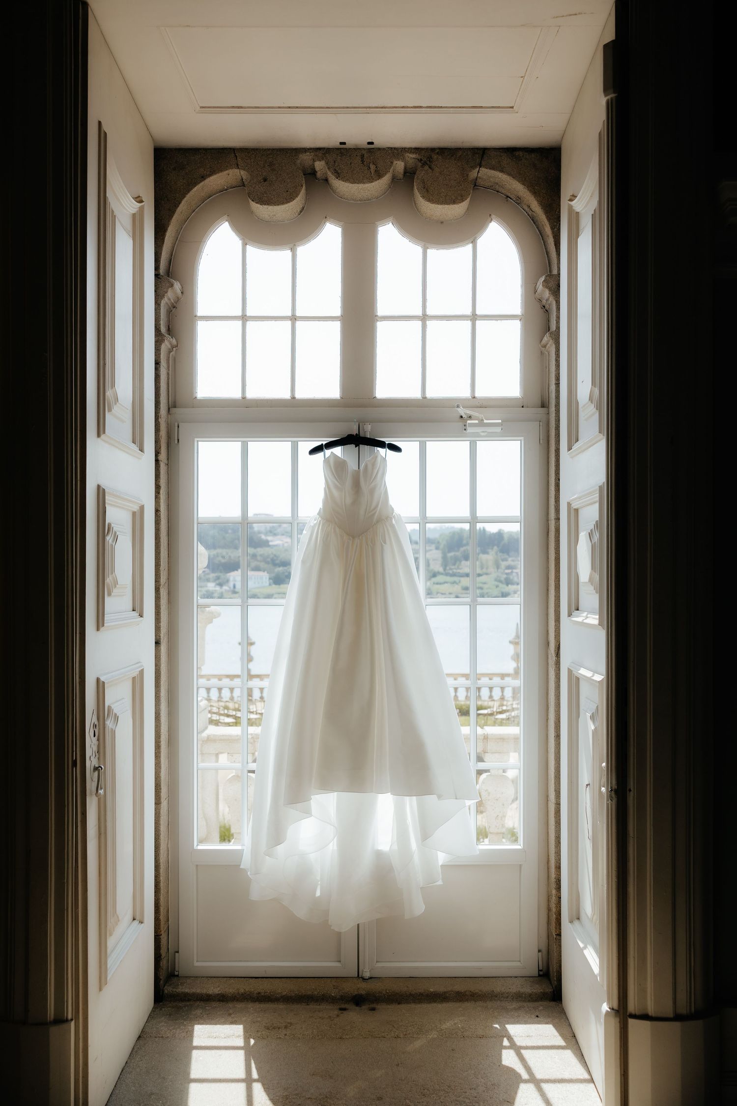 A white wedding dress hangs in a bright window frame overlooking a waterfront view, creating an ethereal silhouette.